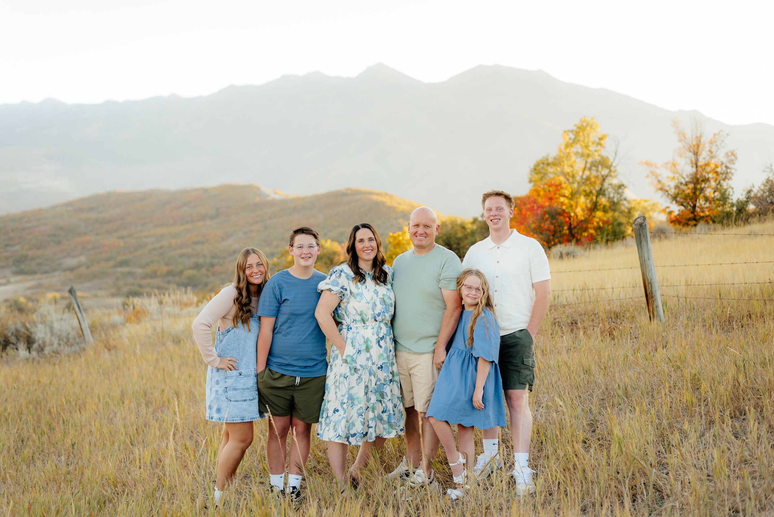 Family of six posing together in a golden field with mountain backdrop and autumn trees during sunset. Megan Snell Photo Utah Weddings