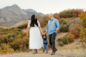 Family of three walking on a mountain trail in autumn, holding hands with toddler between parents. Megan Snell Photo Utah Weddings