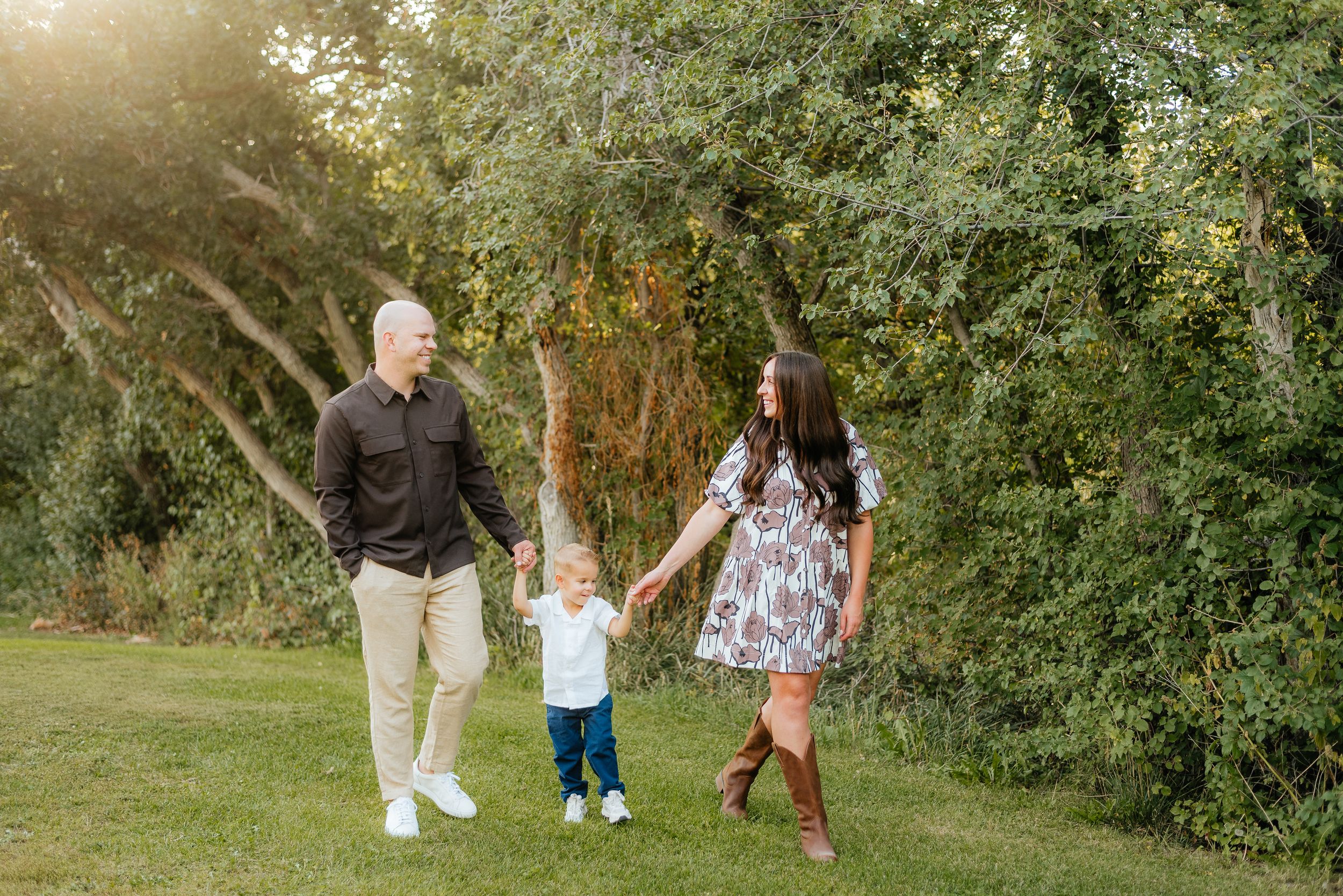 Family of three walking hand-in-hand on green grass with lush trees in golden hour sunlight. Megan Snell Photo Utah Weddings