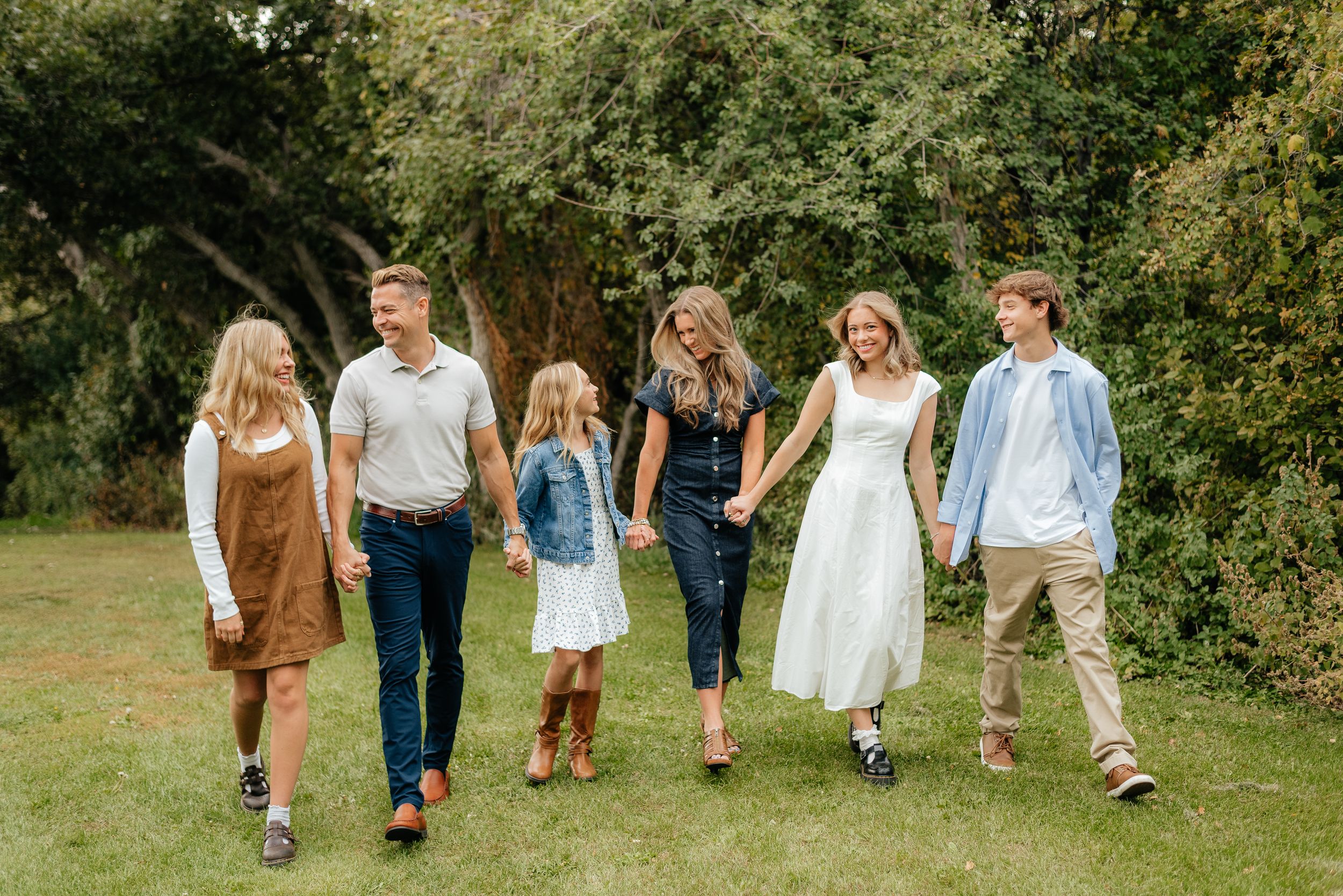 A happy family of six walks hand-in-hand through a lush green park, smiling and enjoying time together outdoors. Megan Snell Photo Utah