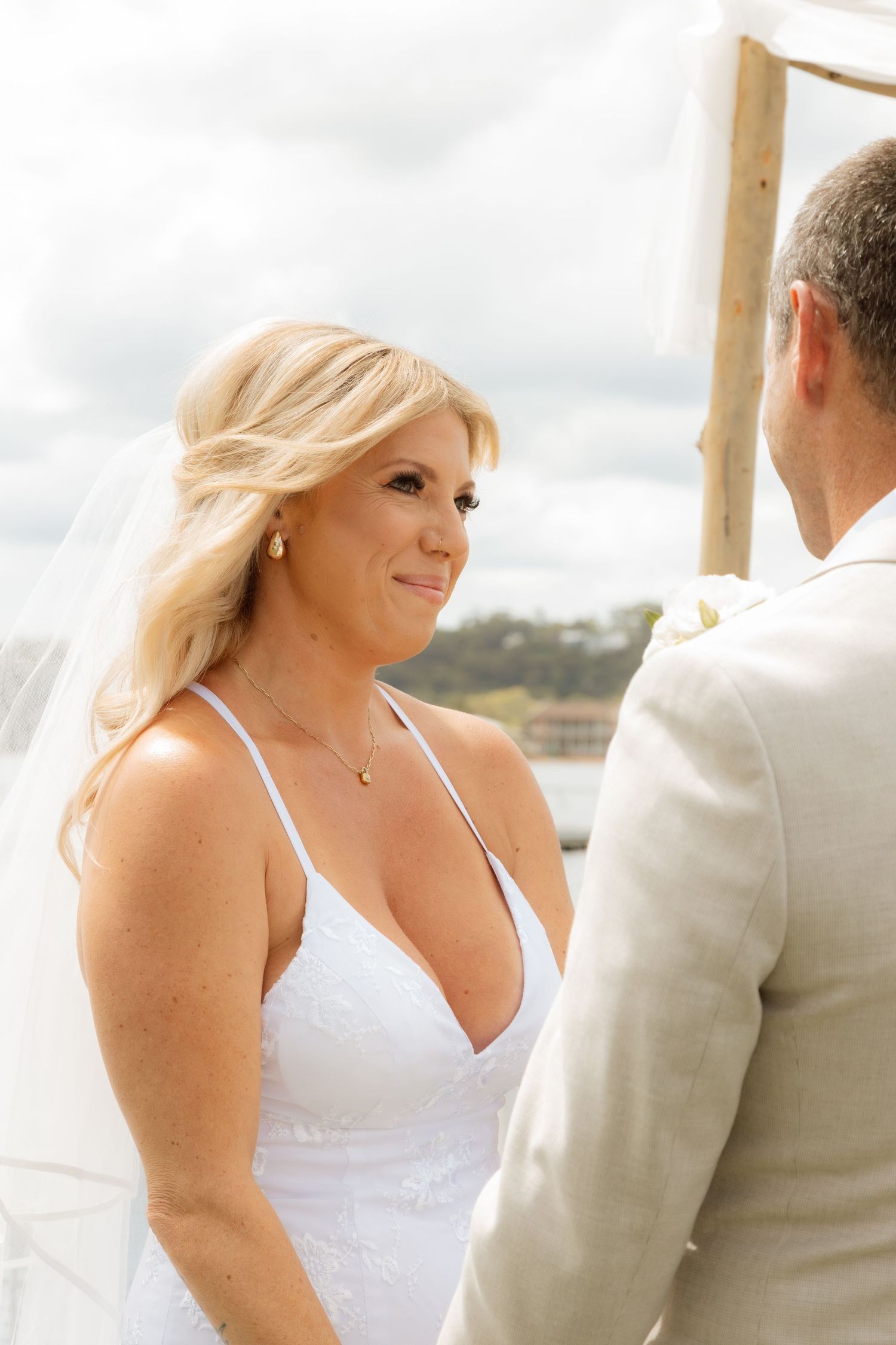 Bride in white halter wedding dress smiles while exchanging vows during outdoor waterfront ceremony.