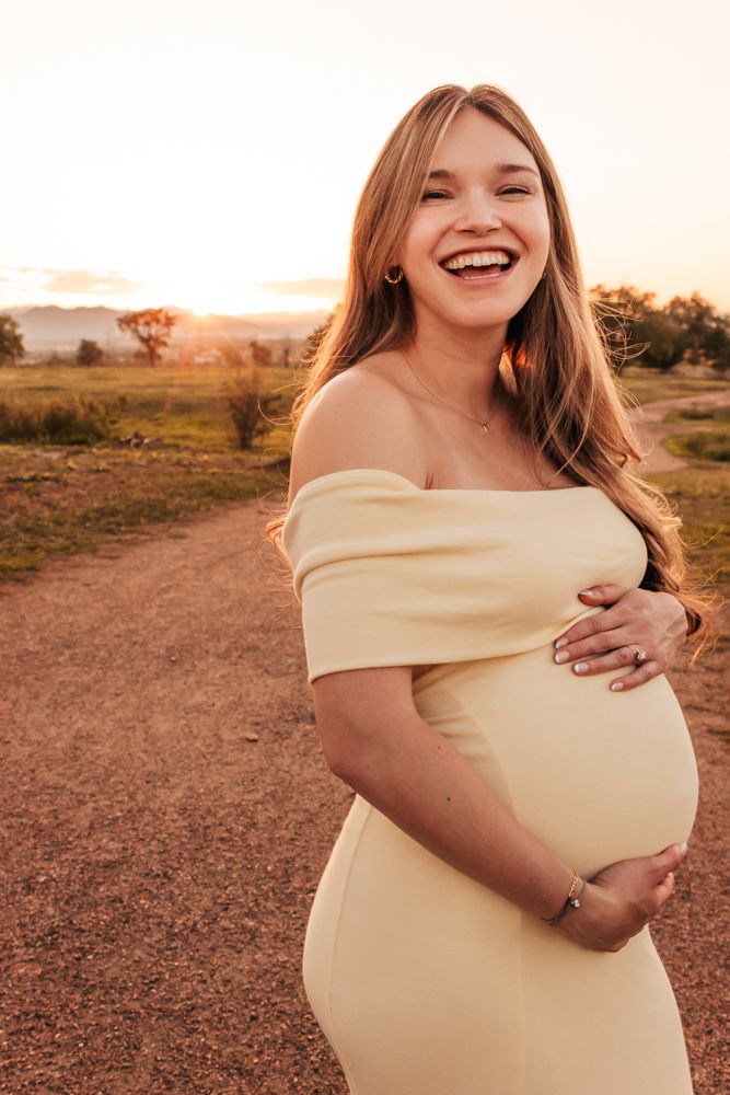 Smiling pregnant woman in yellow off-shoulder dress cradling baby bump at golden hour sunset | Family photographer in  Westminster CO