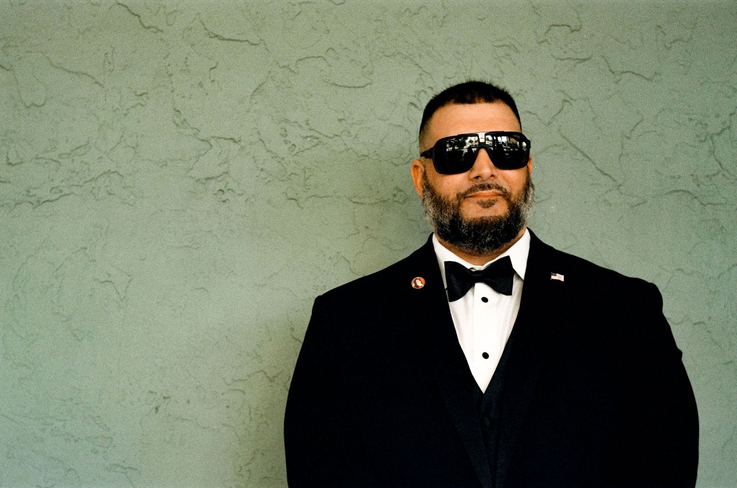 A formal figure in a black tuxedo and sunglasses stands against a textured green wall.