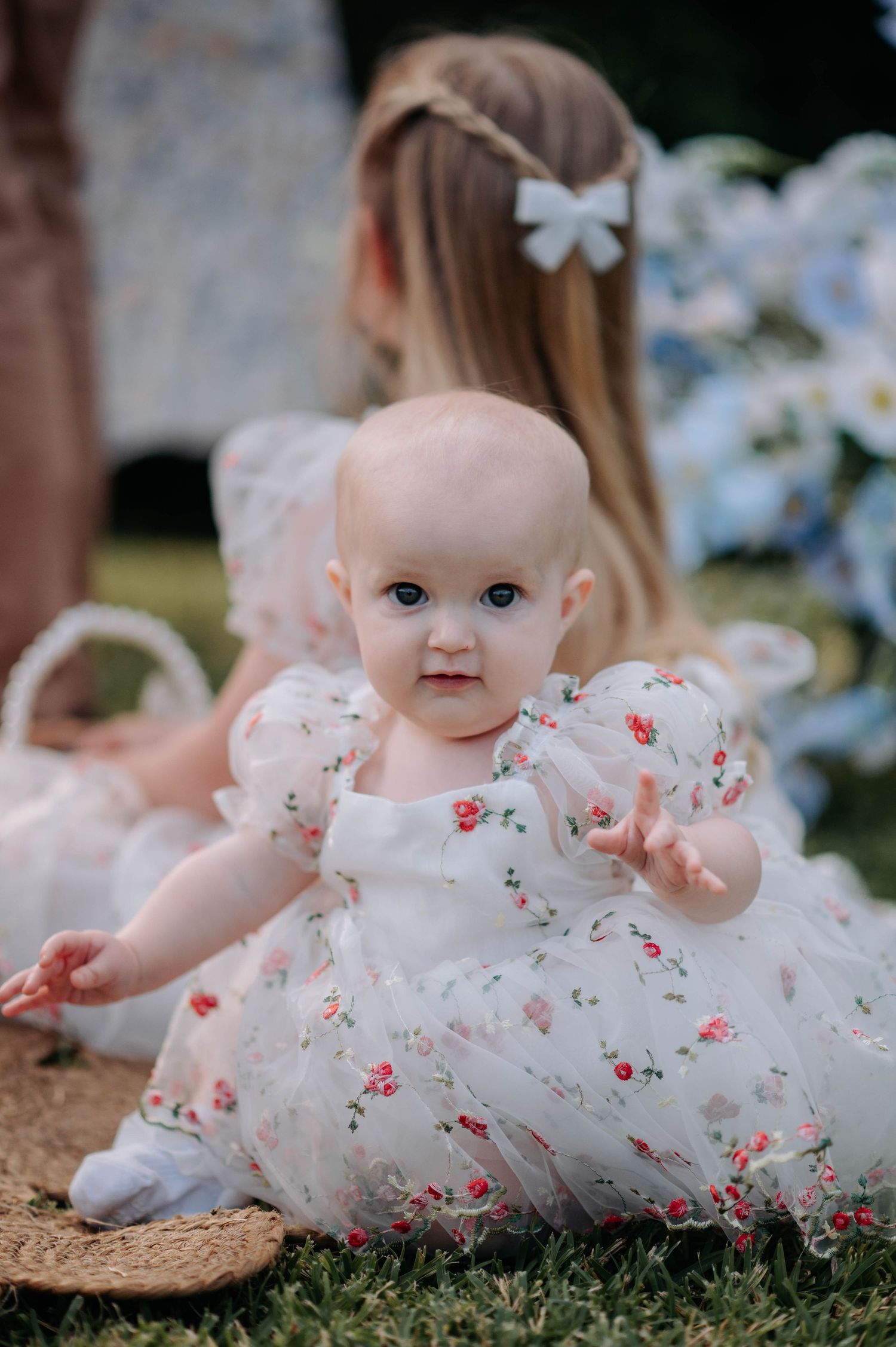 Baby in white floral dress with bow sitting on ground with flowers in background.