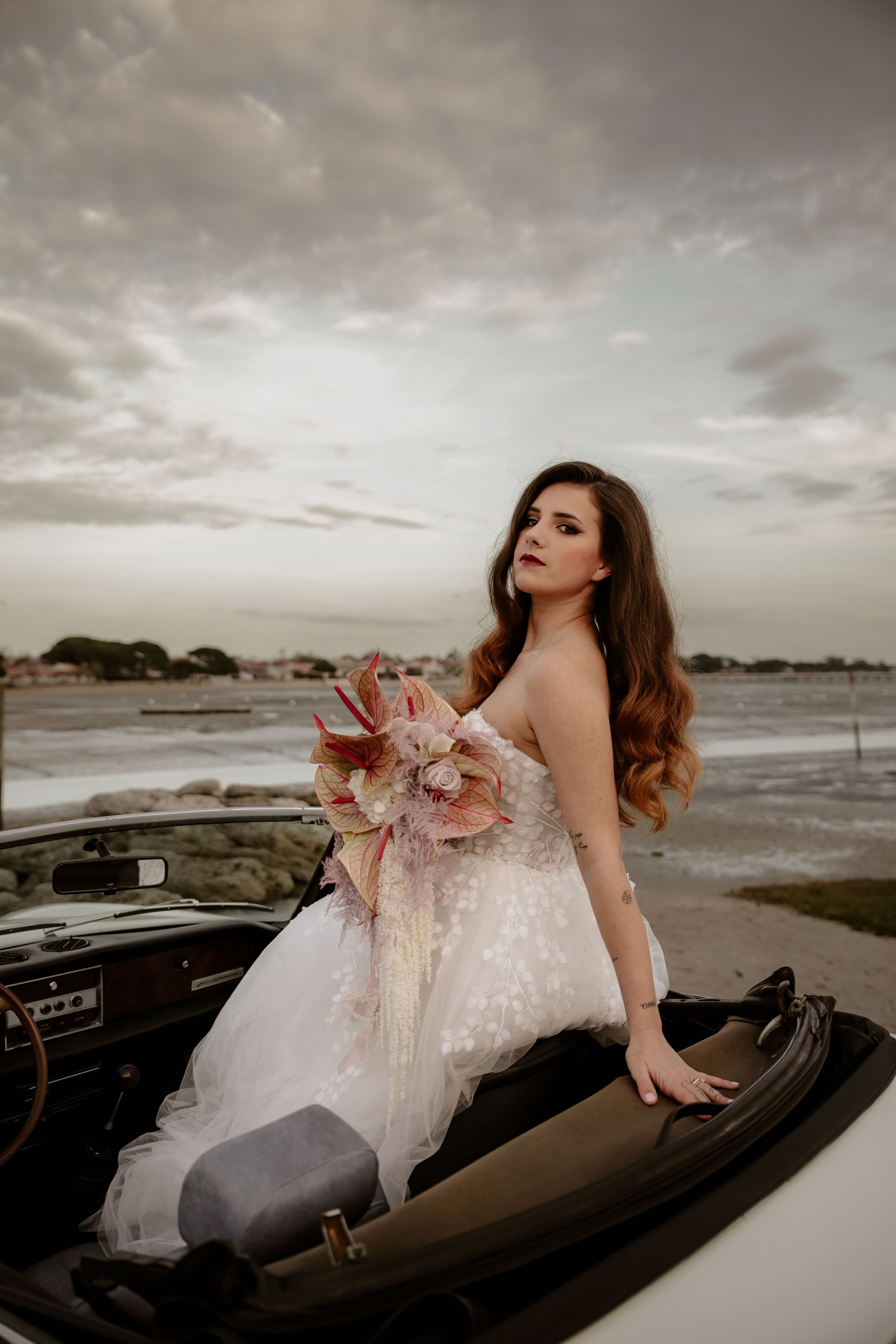 Bride in lace gown poses with pink floral bouquet in white classic car by the ocean.
