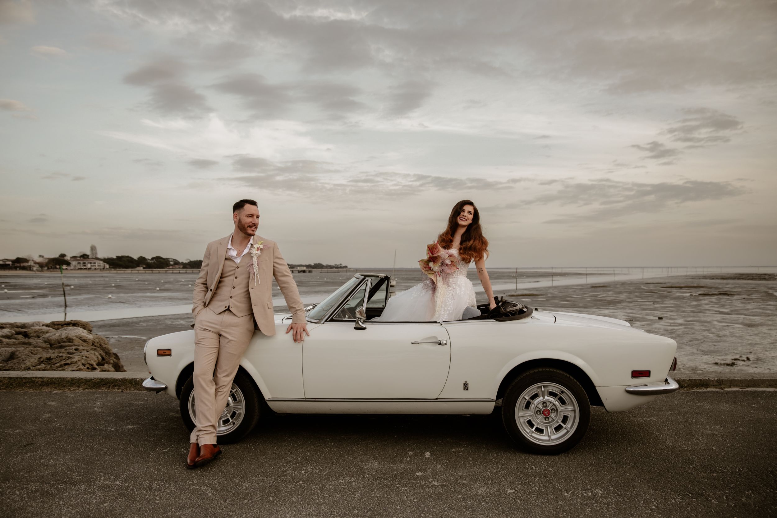 Newlywed couple poses with vintage white convertible car on coastal road during sunset.