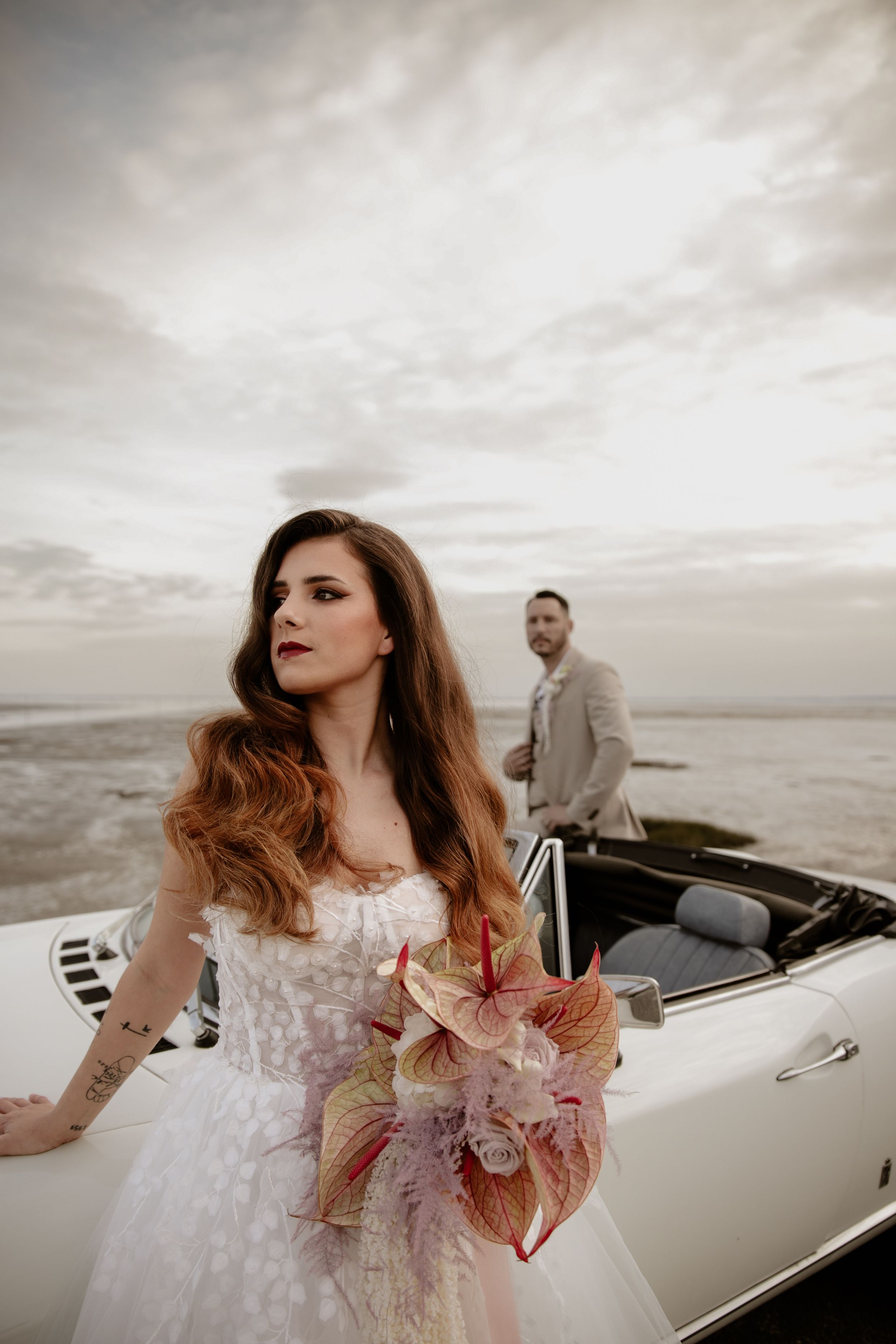 A bride in a white dress holds a pink bouquet while posing in front of a classic convertible car at sunset.