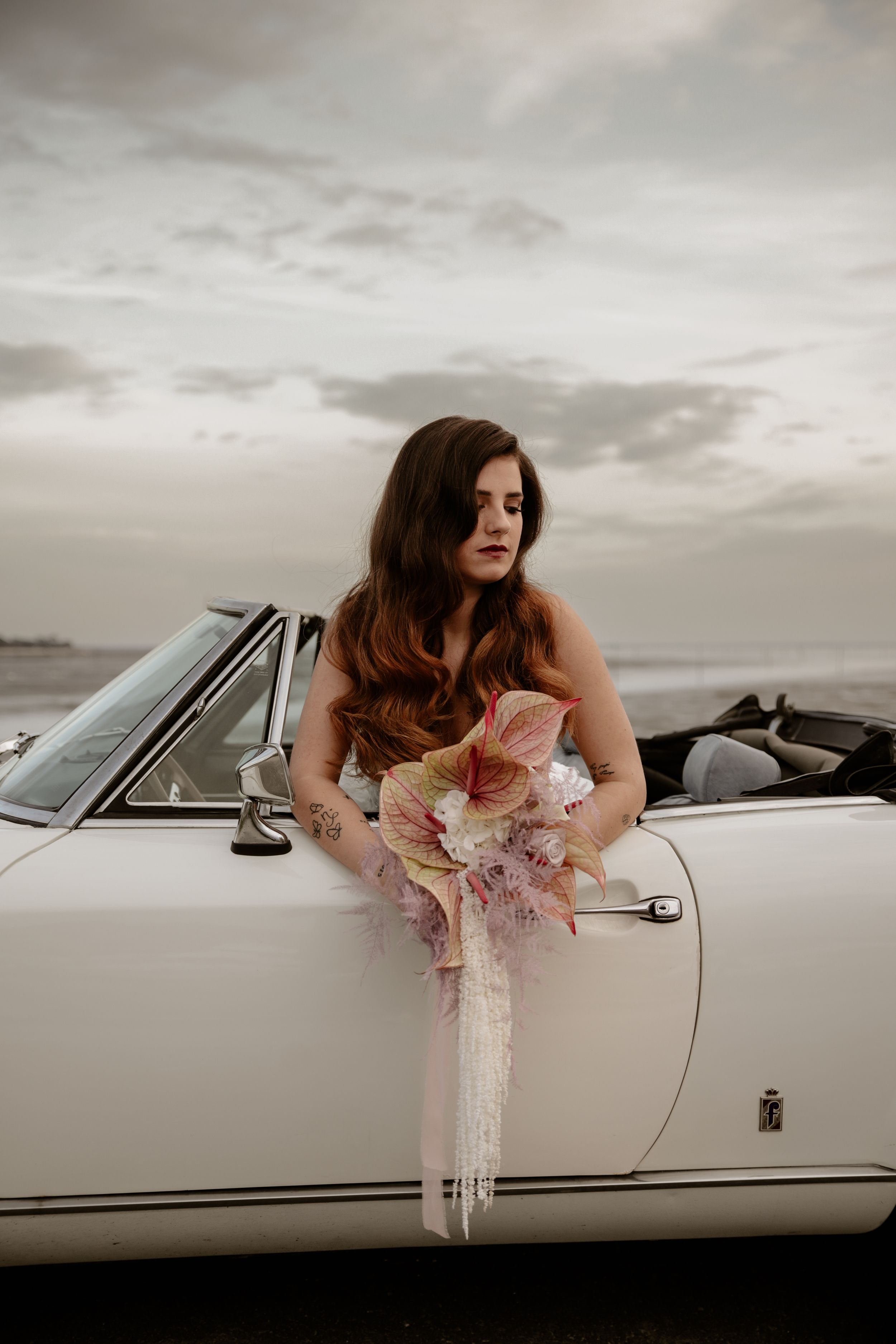 Elegant bride with long wavy hair leans against classic white convertible holding pink flowers.