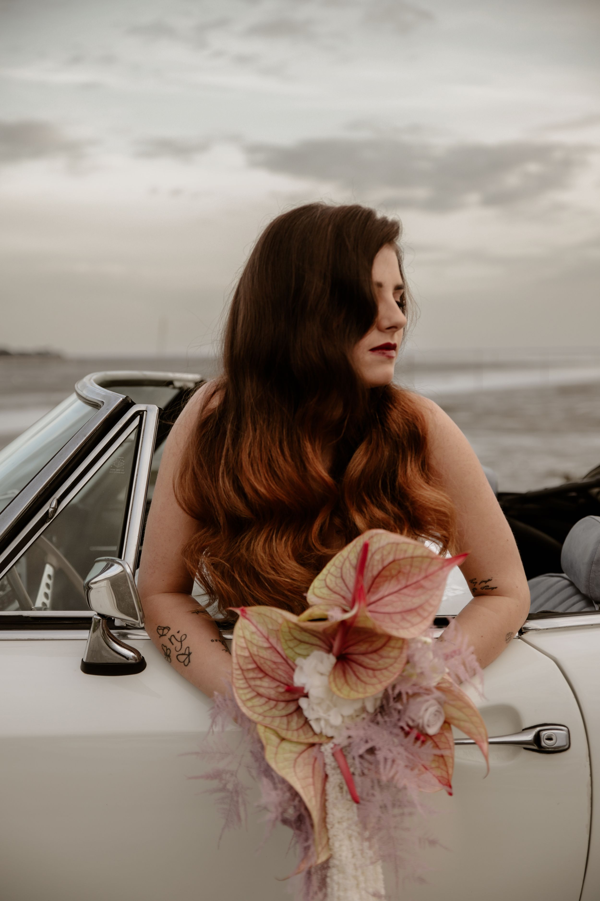 Bride with flowing dark hair poses with antique flowers against vintage car door.