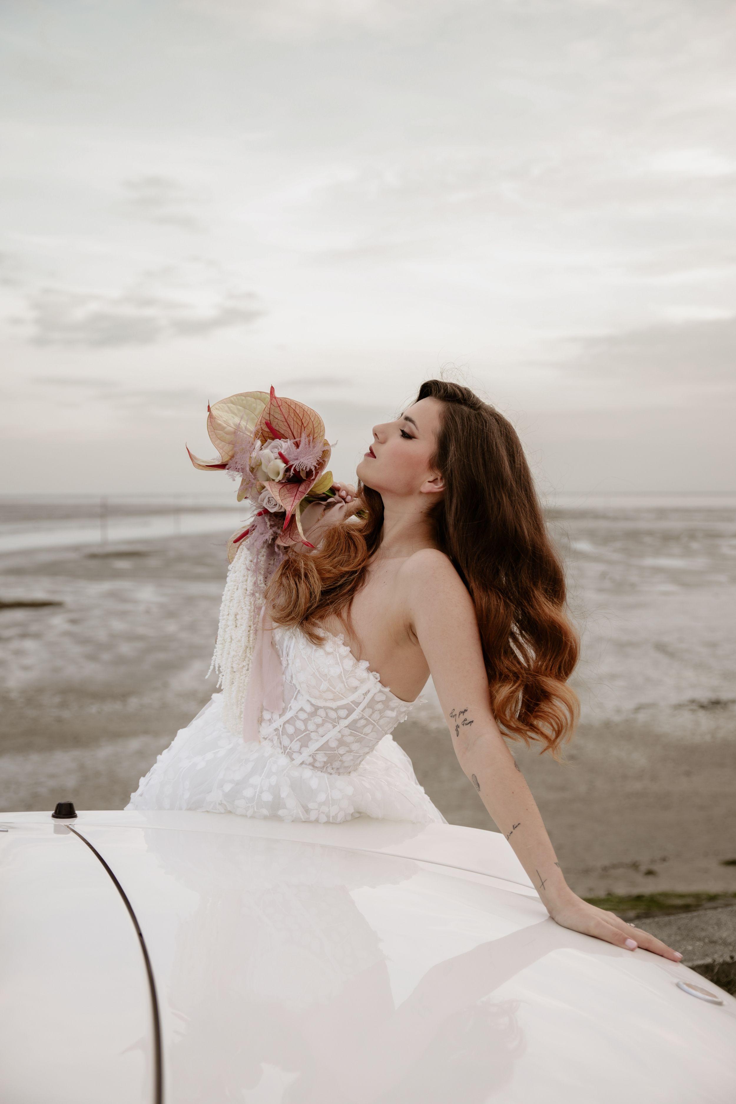 Bride in white dress poses dramatically on classic car hood with pink bouquet by ocean.