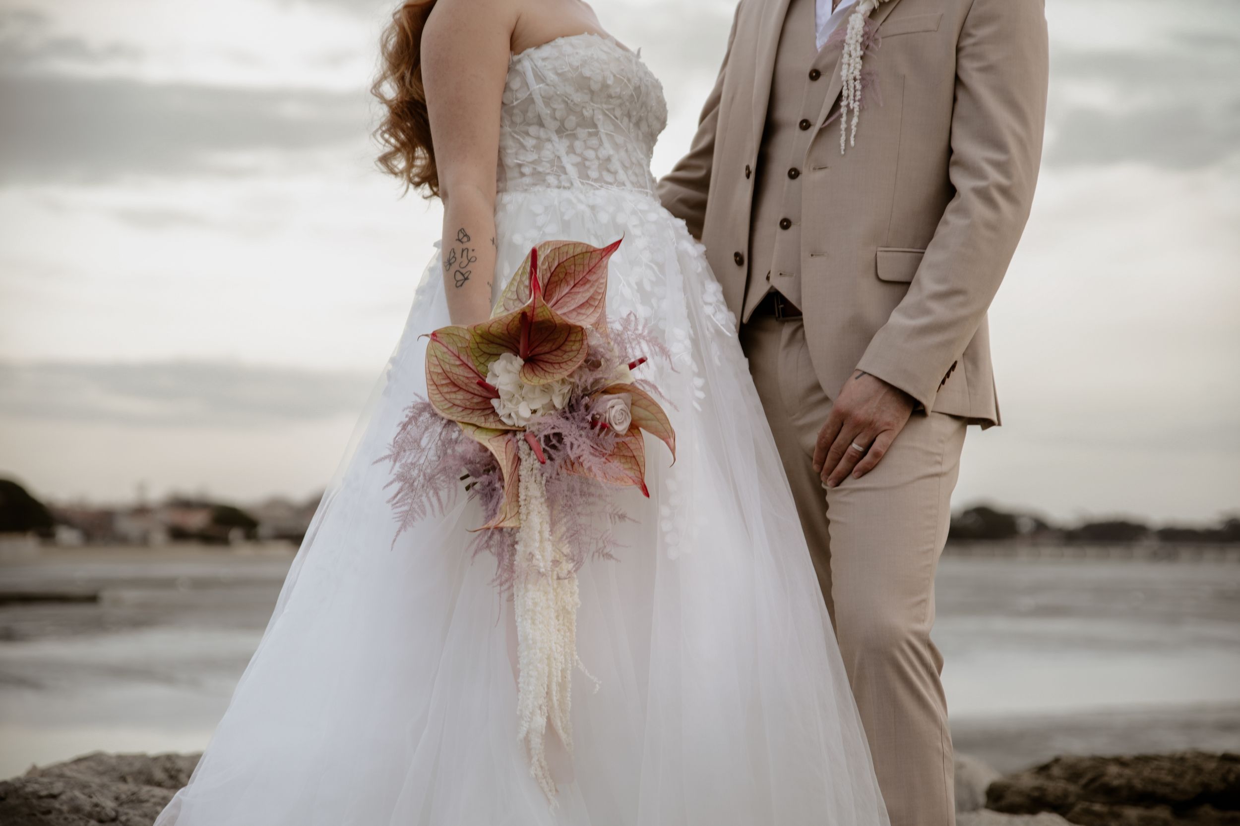 Couple in wedding attire standing by ocean with bride holding pink and cream bouquet.