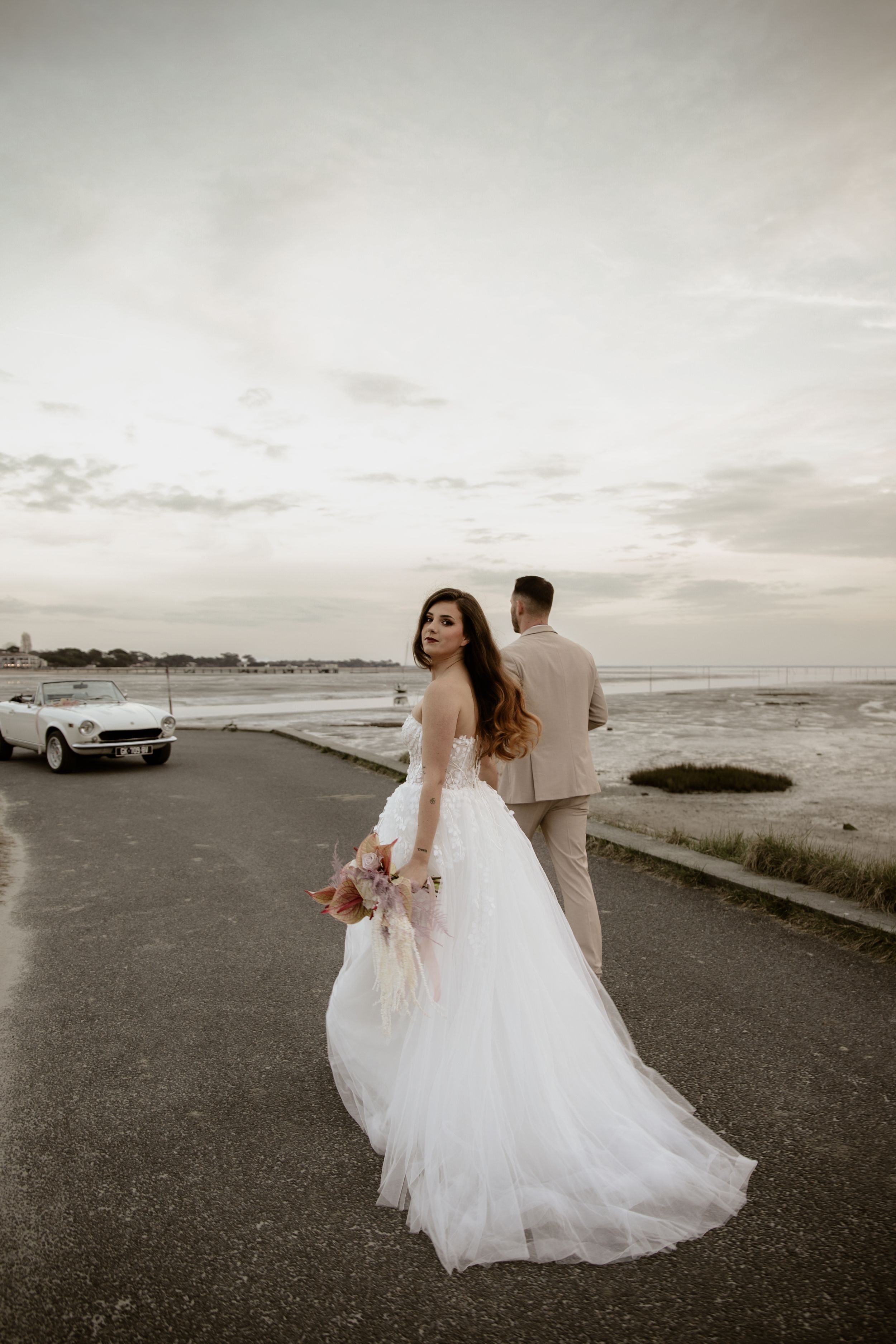 Bride in flowing white gown and groom in tan suit pose on coastal road with vintage car.