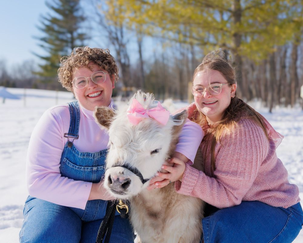 Two people in pink sweaters pose outdoors with a white miniature pony with a pink bow in its mane on a snowy winter day.
