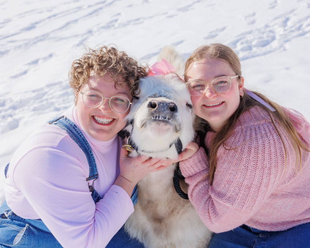 Two people in winter clothing pose outdoors in the snow with a fluffy white dog between them.