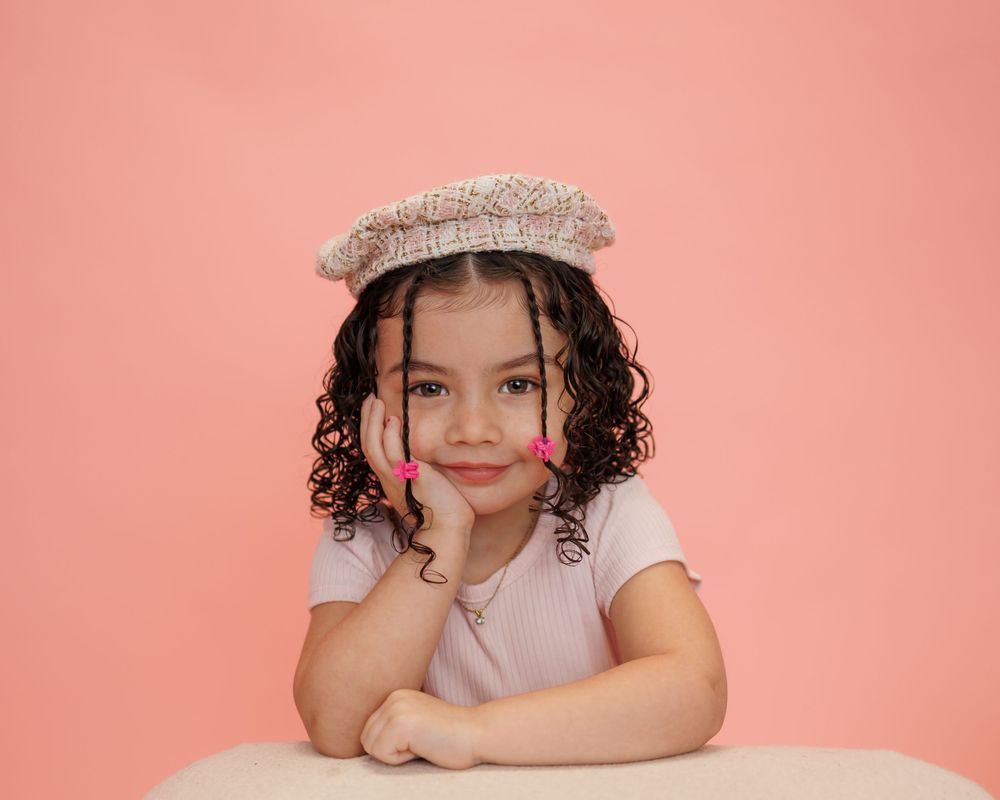 A child wearing a beaded hair accessory poses against a pink background with curly hair and white shirt.