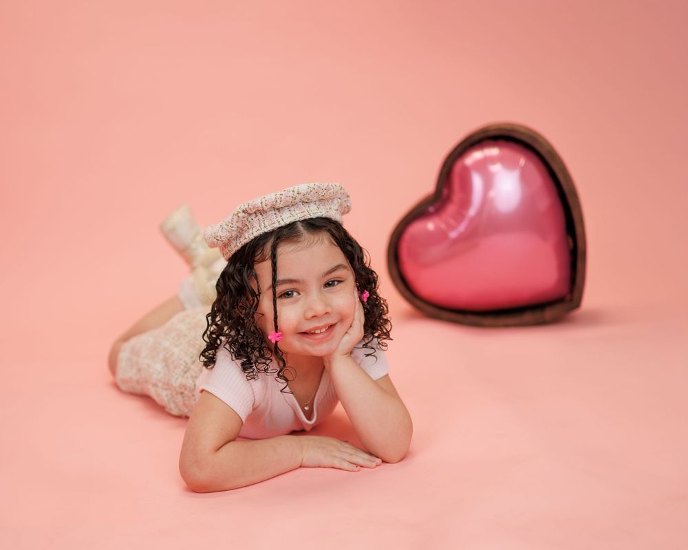 Sweet portrait of a young child laying on a pink backdrop with a large red heart decoration.