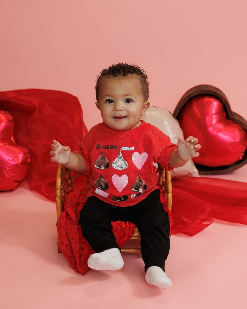 An infant sits on a pink background surrounded by red heart decorations and wearing a festive Valentine's Day outfit.