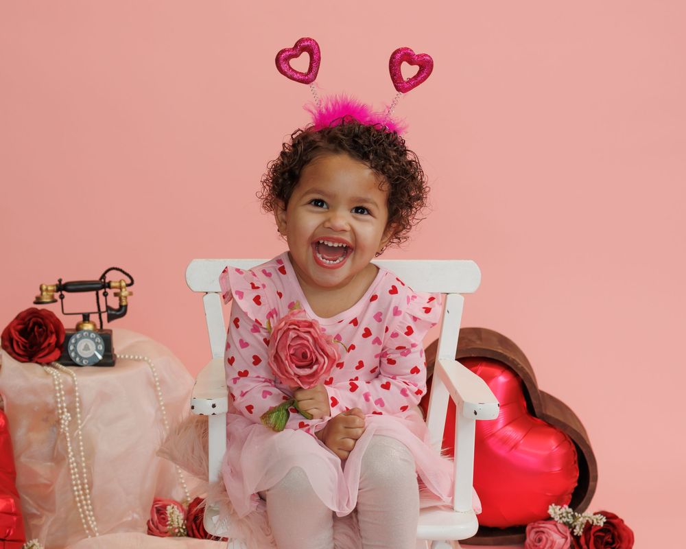 Child wearing pink valentine outfit sits on white chair with heart decor against pink backdrop, holding rose and smiling.