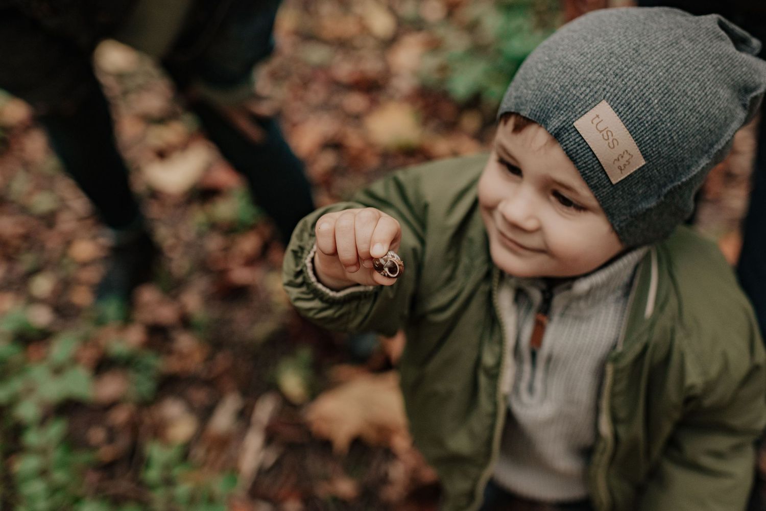 A group of children in green jackets and winter hats explore autumn leaves in a forest setting.