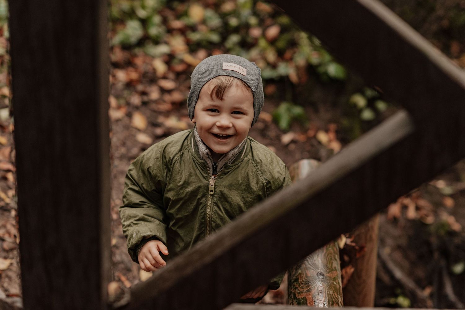 A person in camouflage clothing smiles while sitting in a wooded outdoor setting with fallen autumn leaves.