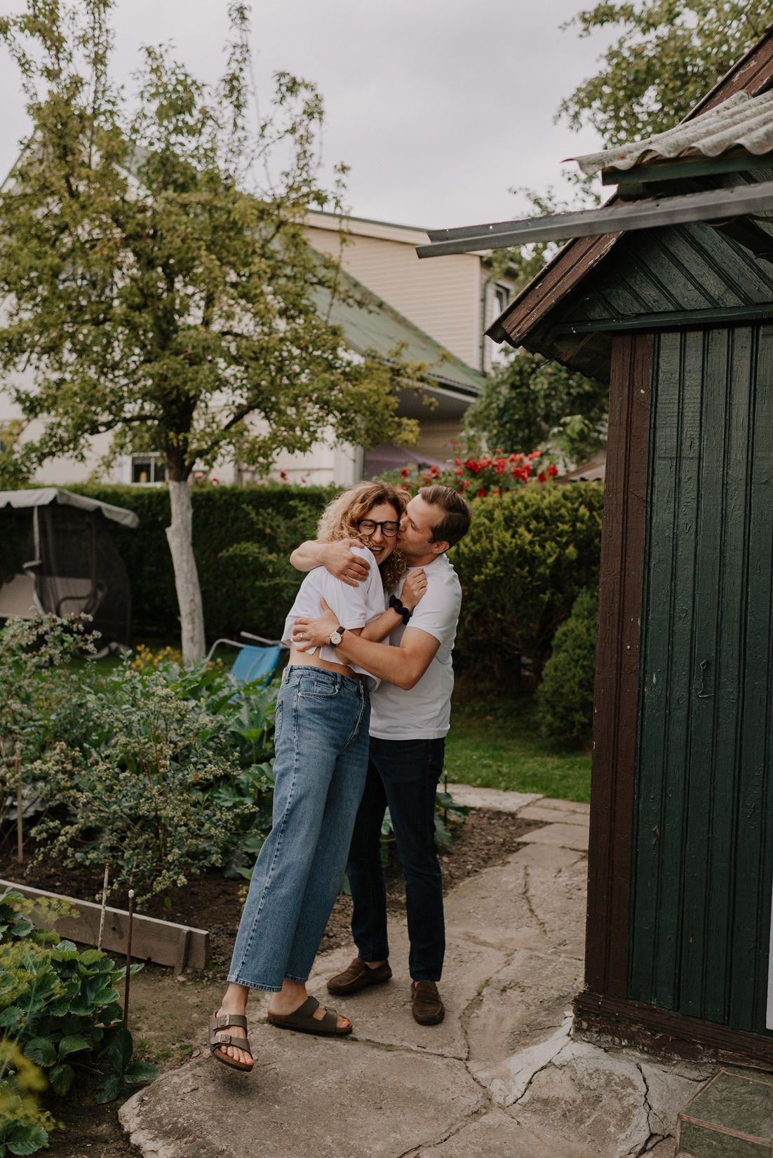 A couple shares an intimate moment together in a garden setting with white buildings and greenery.