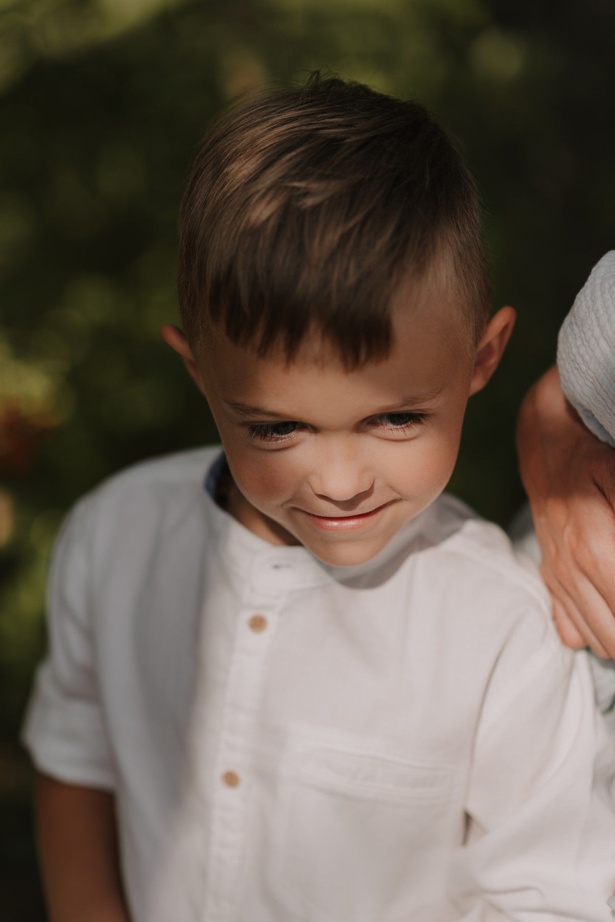 A close-up portrait showing a soft smile in a white shirt against a natural outdoor backdrop.