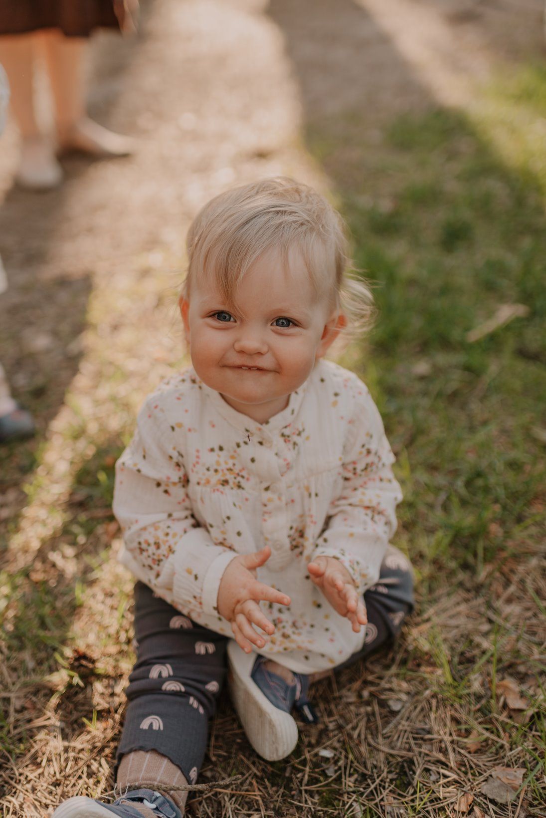 Small child in patterned white top sits on grass during golden hour photography session.