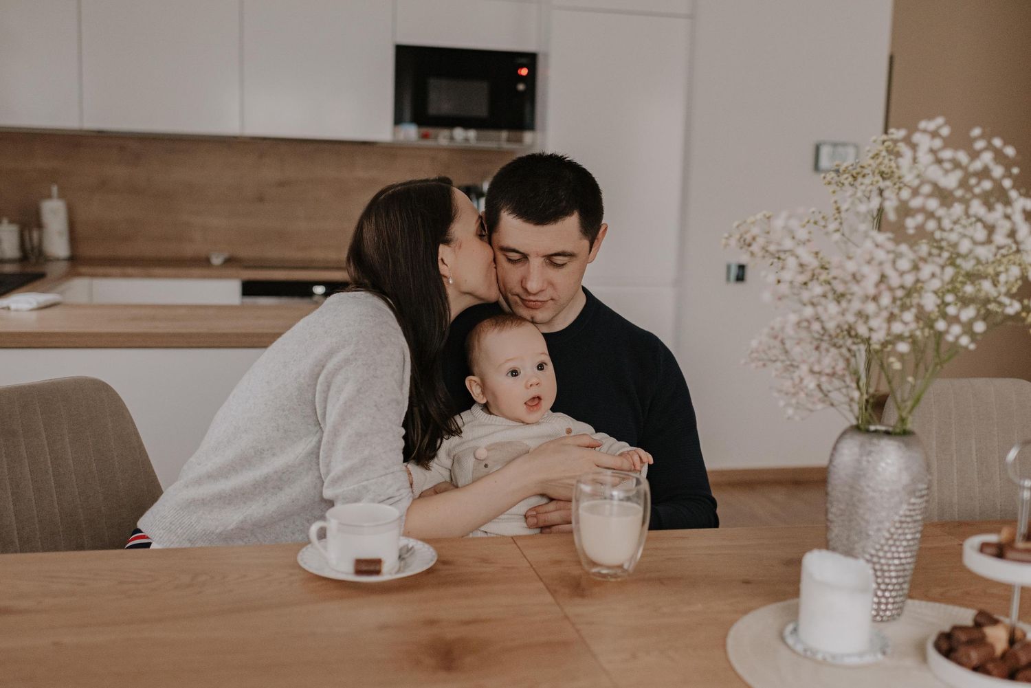 Family sharing an intimate moment around a wooden dining table with baby, flowers and coffee cups.