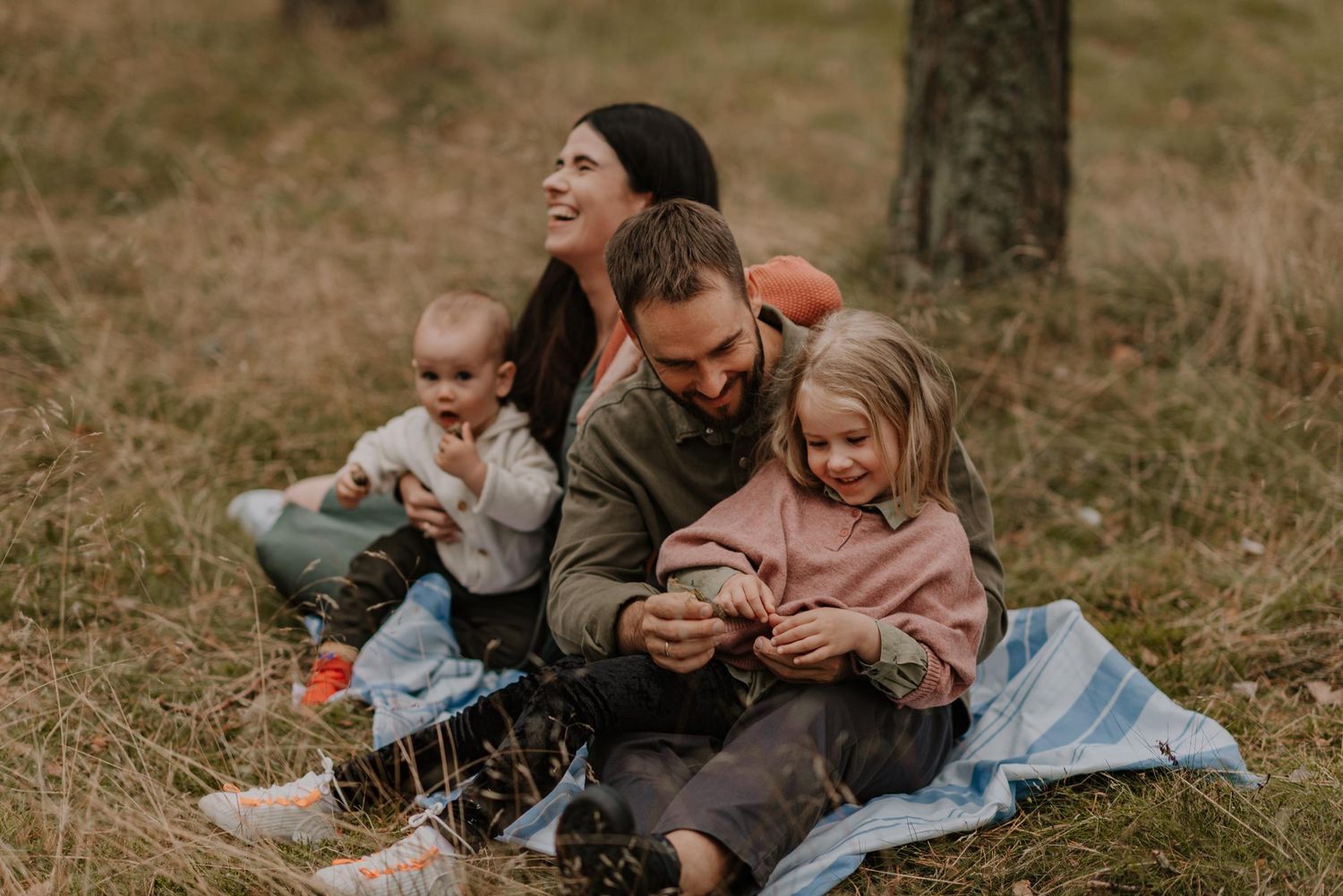 Family relaxing together on picnic blanket in wooded setting.