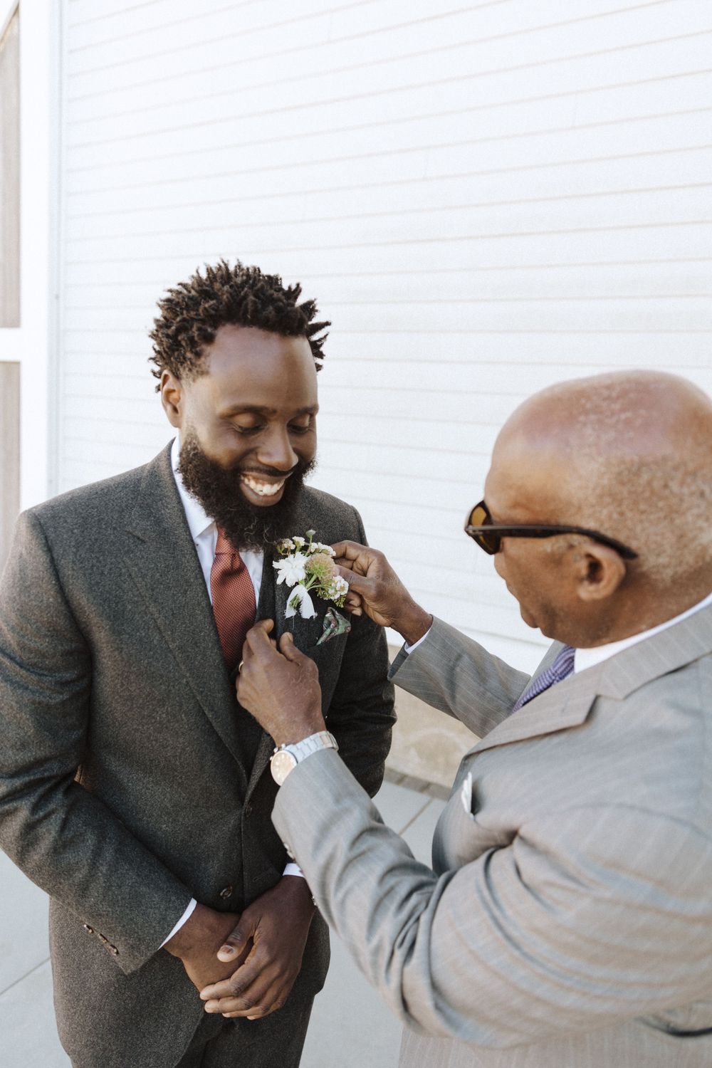 Man in gray suit smiles warmly while someone adjusts his white floral boutonniere before formal event in sussex county nj.