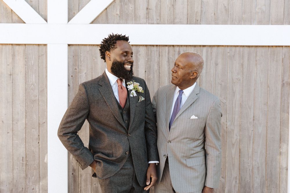 Two grooms in stylish suits share a joyful moment together against a white and gray barnwood wall at their wedding in sussex county nj.