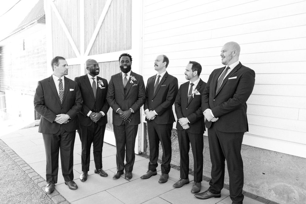 Black and white photo of six groomsmen in dark suits standing together and smiling against a white wall in sussex county nj.