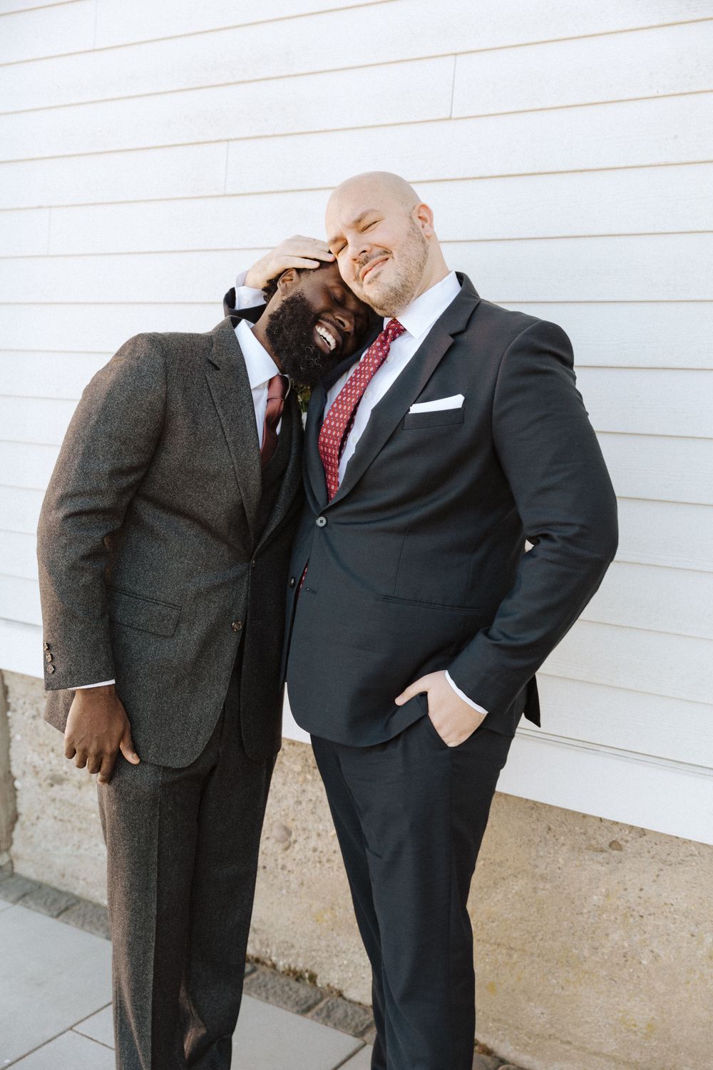 Two men in suits sharing a casual, lighthearted moment while standing against a white wall on a farm in vernon nj.