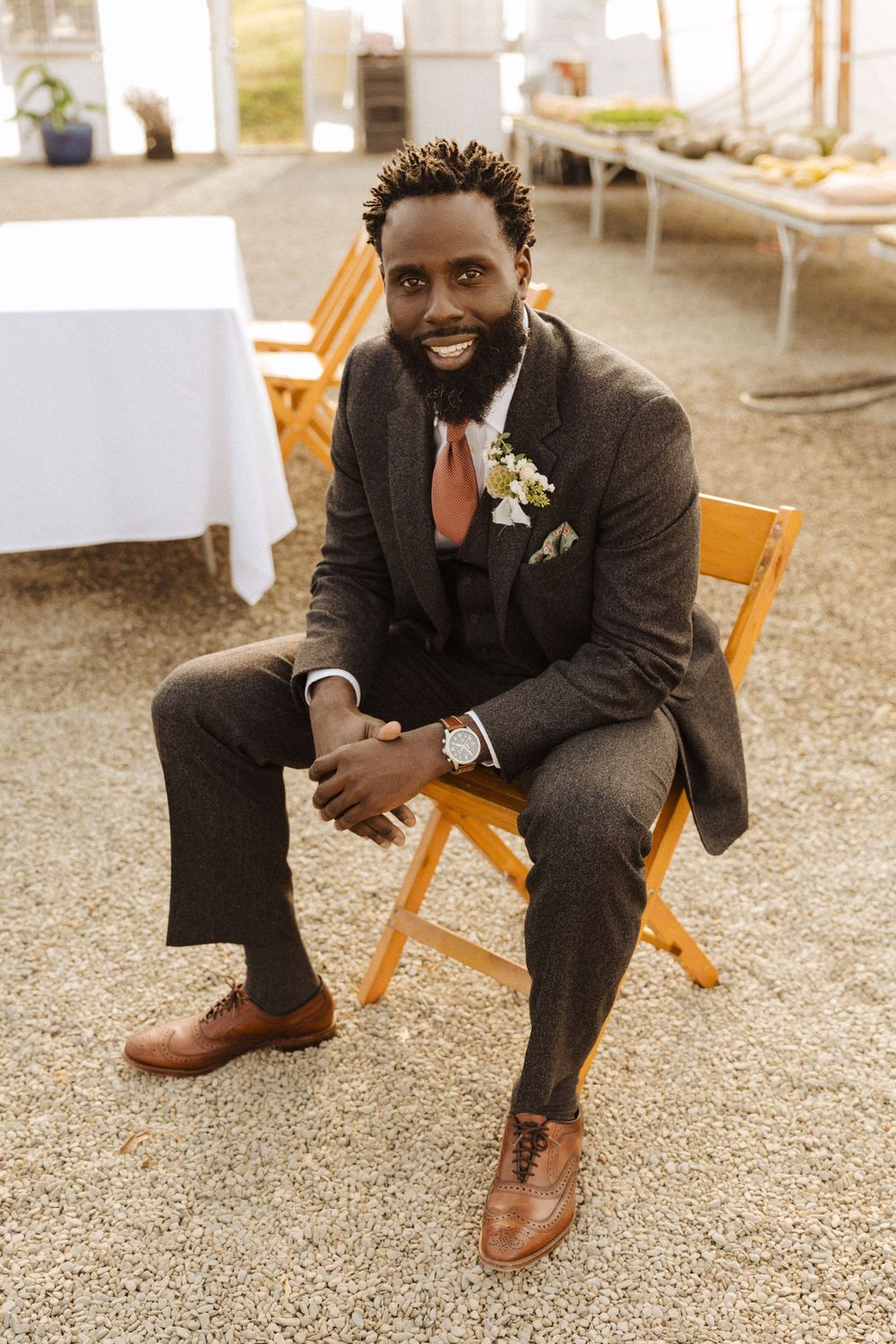 Wedding guest in dark suit and red tie sits smiling on wooden folding chair at outdoor reception venue in vernon nj.