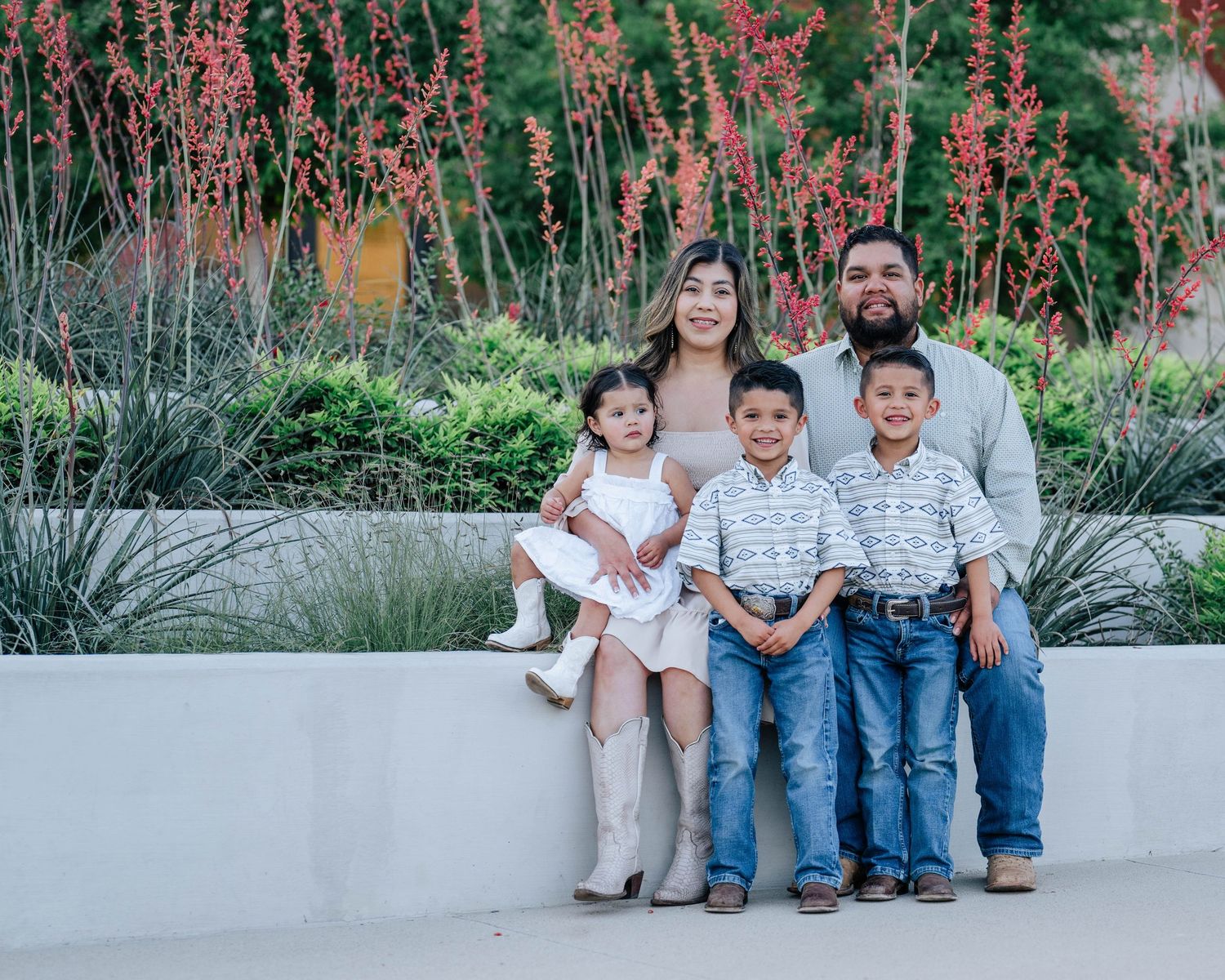 Family of five sits together on concrete bench with flowering plants in background.