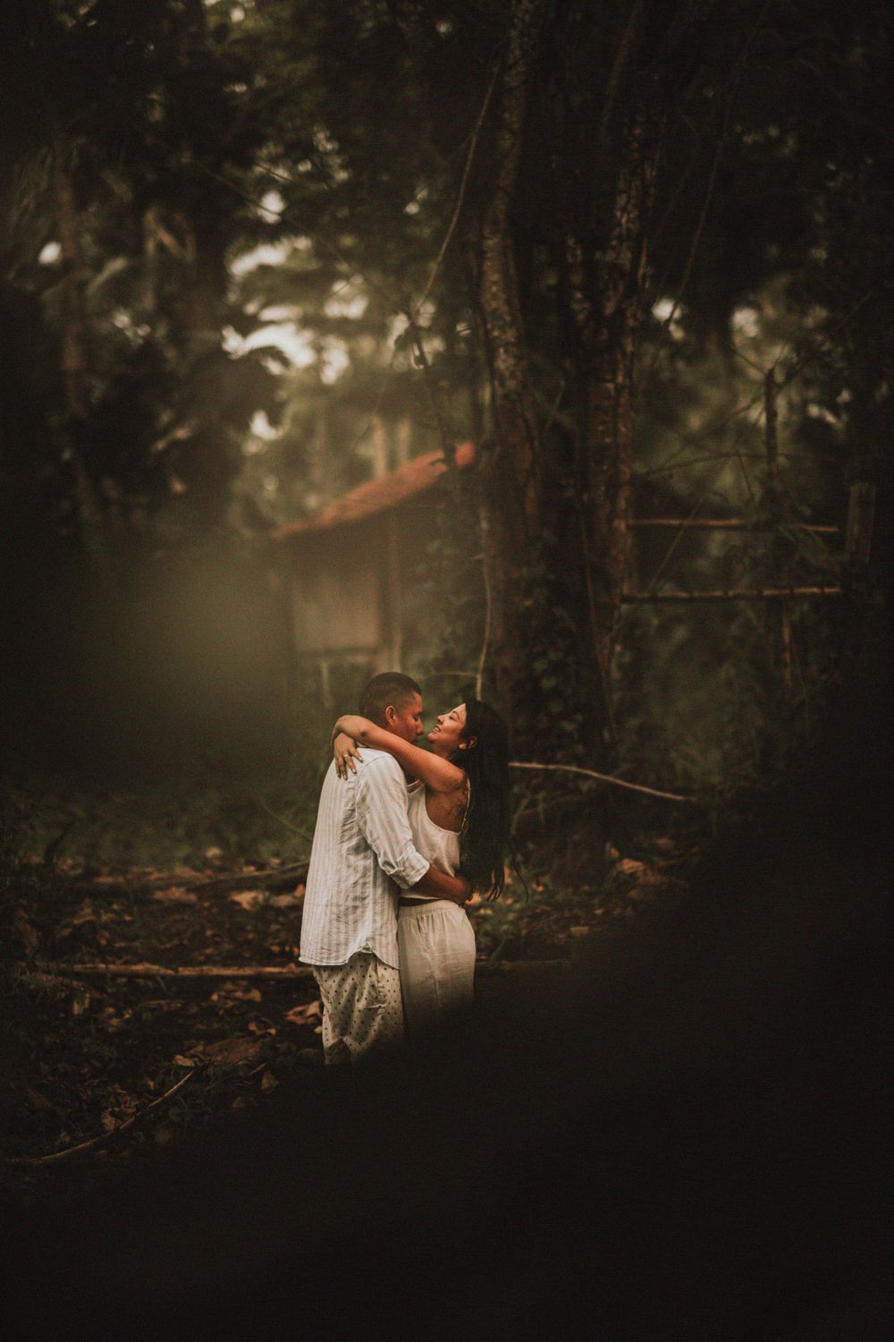 A couple embraces in a moody forest setting with dark shadows and filtered sunlight.