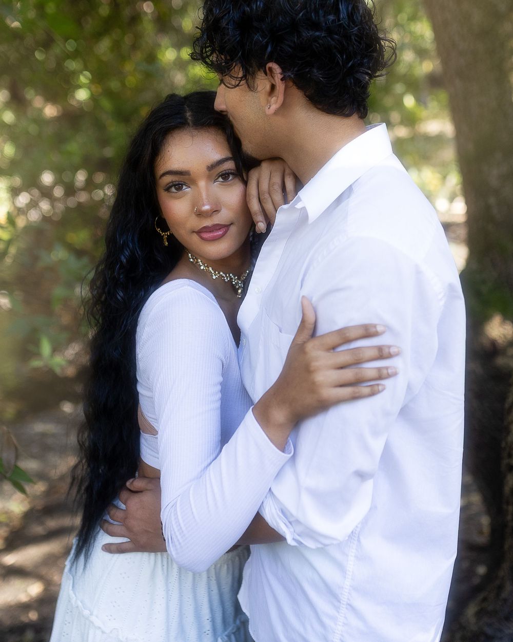 A romantic couple in white clothing embraces tenderly in a sun-dappled outdoor setting.