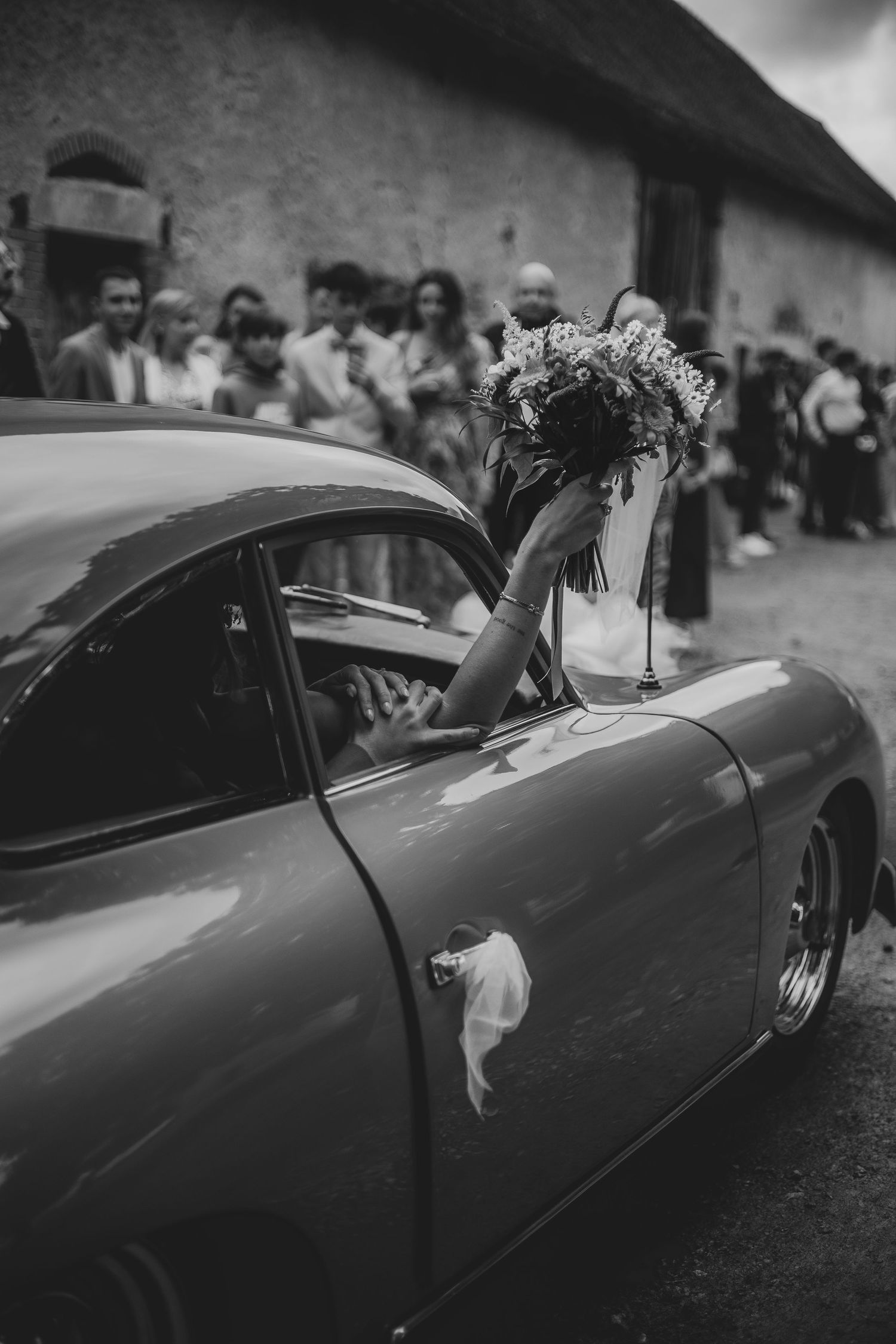 Photo en noir et blanc d'une voiture ancienne avec une mariée à l'intérieur tenant la main de son partenaire.
