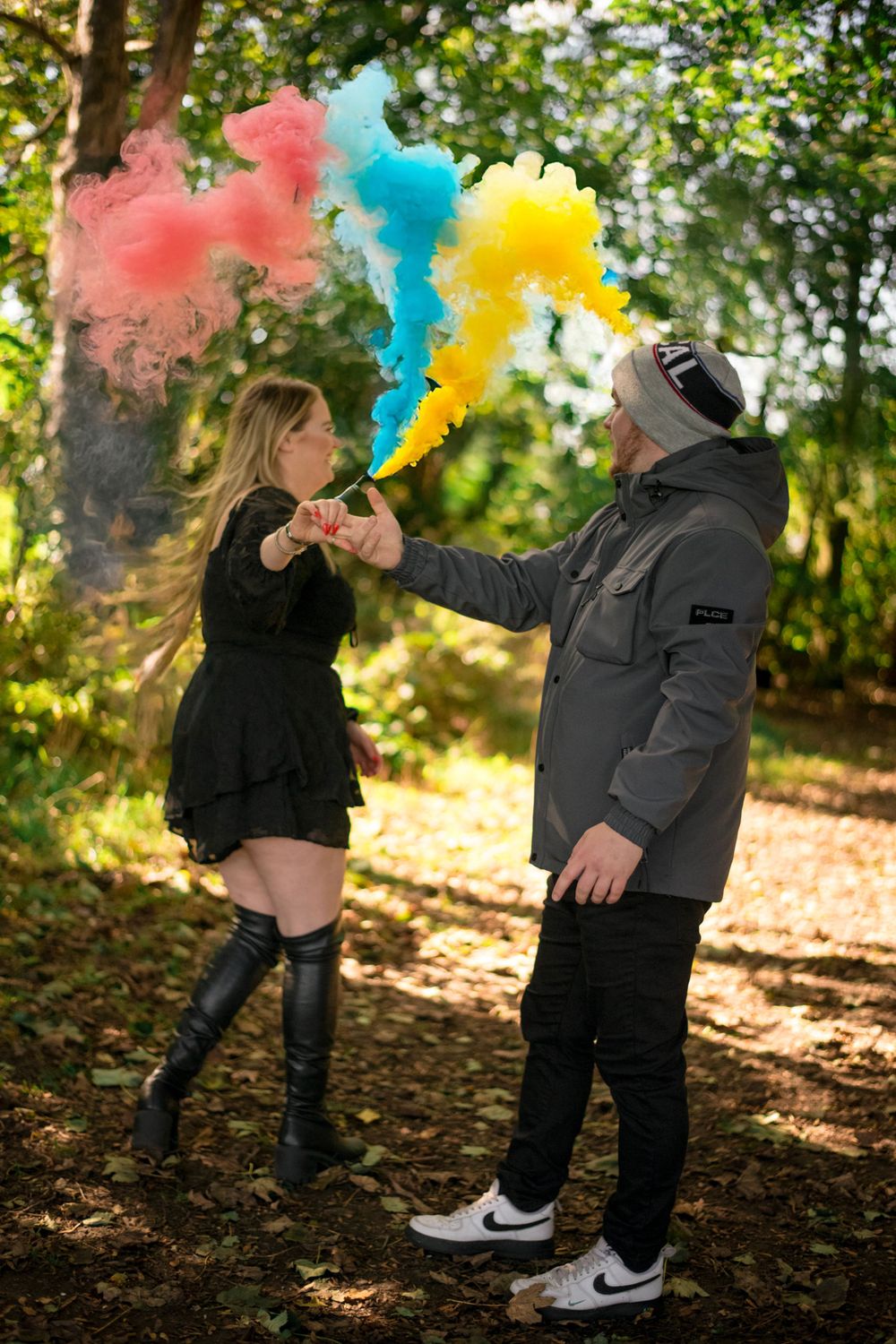 A couple plays with colorful smoke bombs in an autumn forest setting creating vibrant red, blue and yellow clouds.