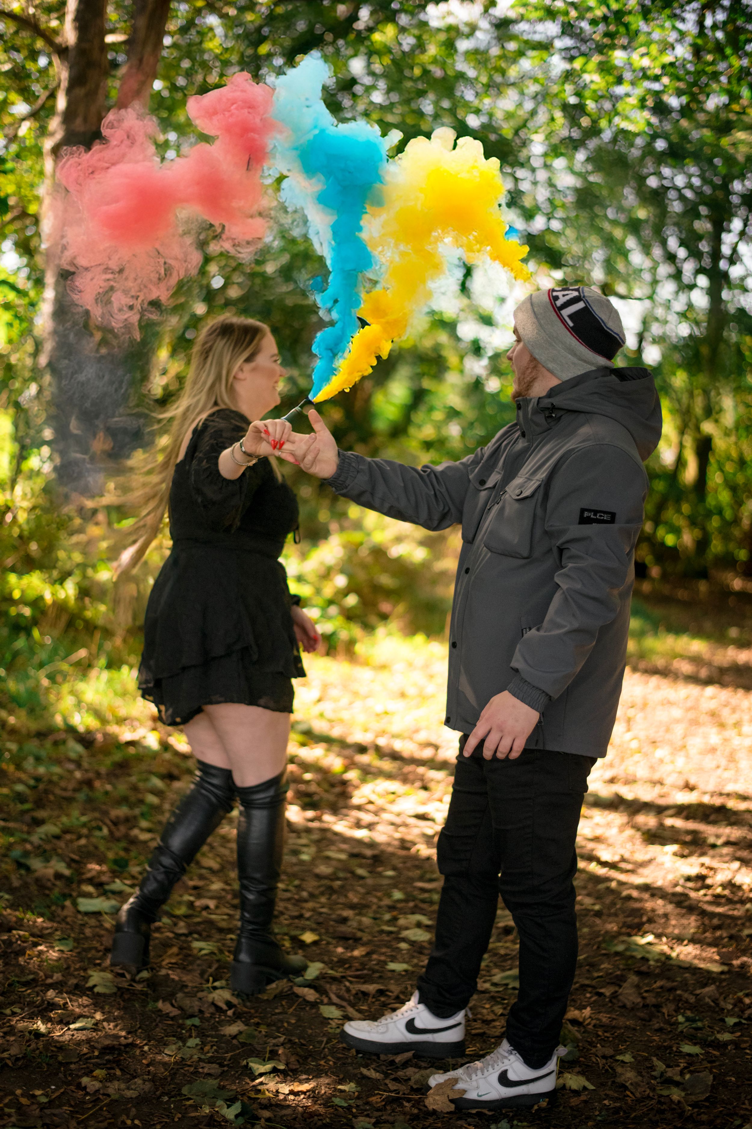 A couple plays with colorful smoke bombs in an autumn forest setting creating vibrant red, blue and yellow clouds.