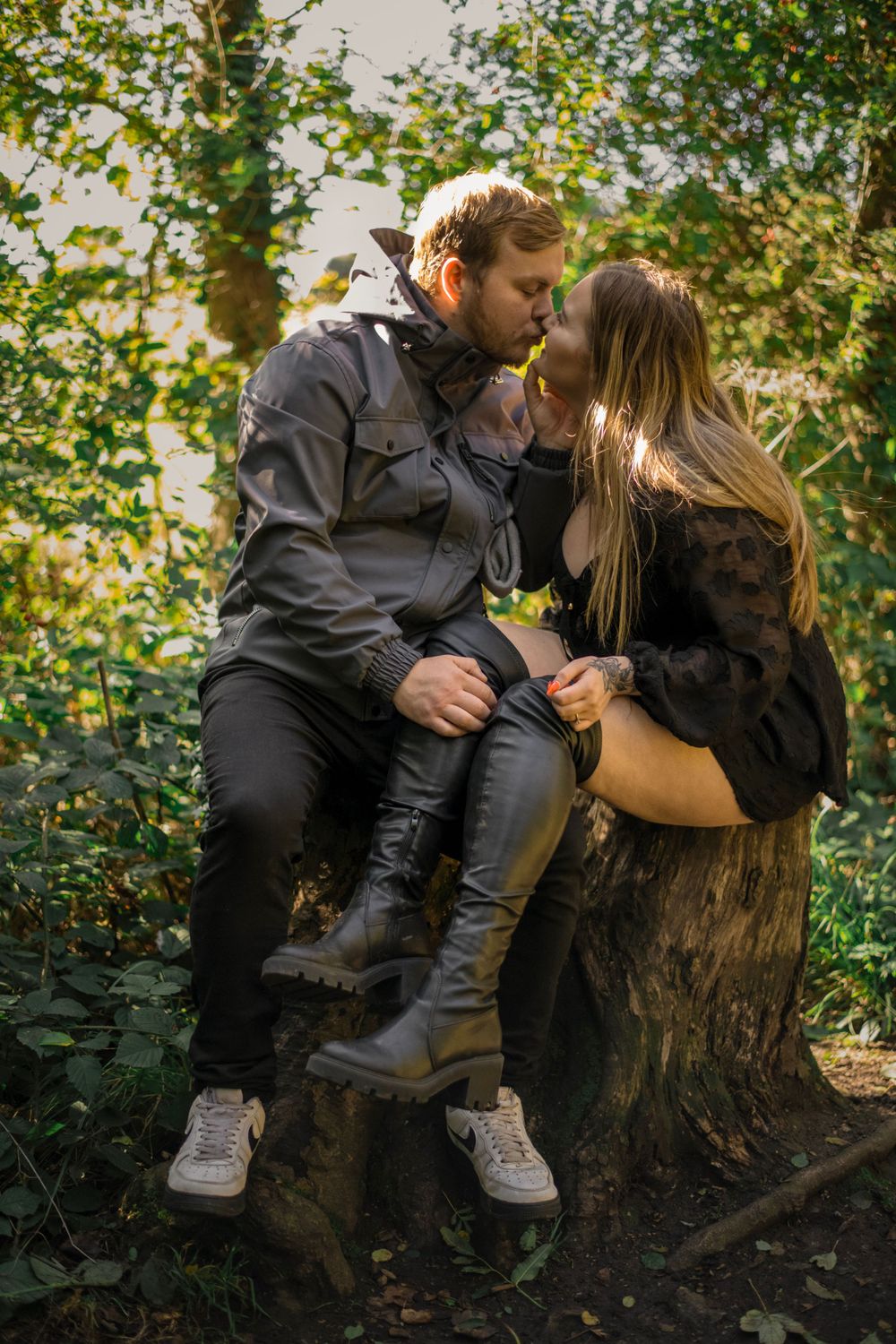 A couple shares an intimate moment while sitting on a tree stump in a sunlit forest.