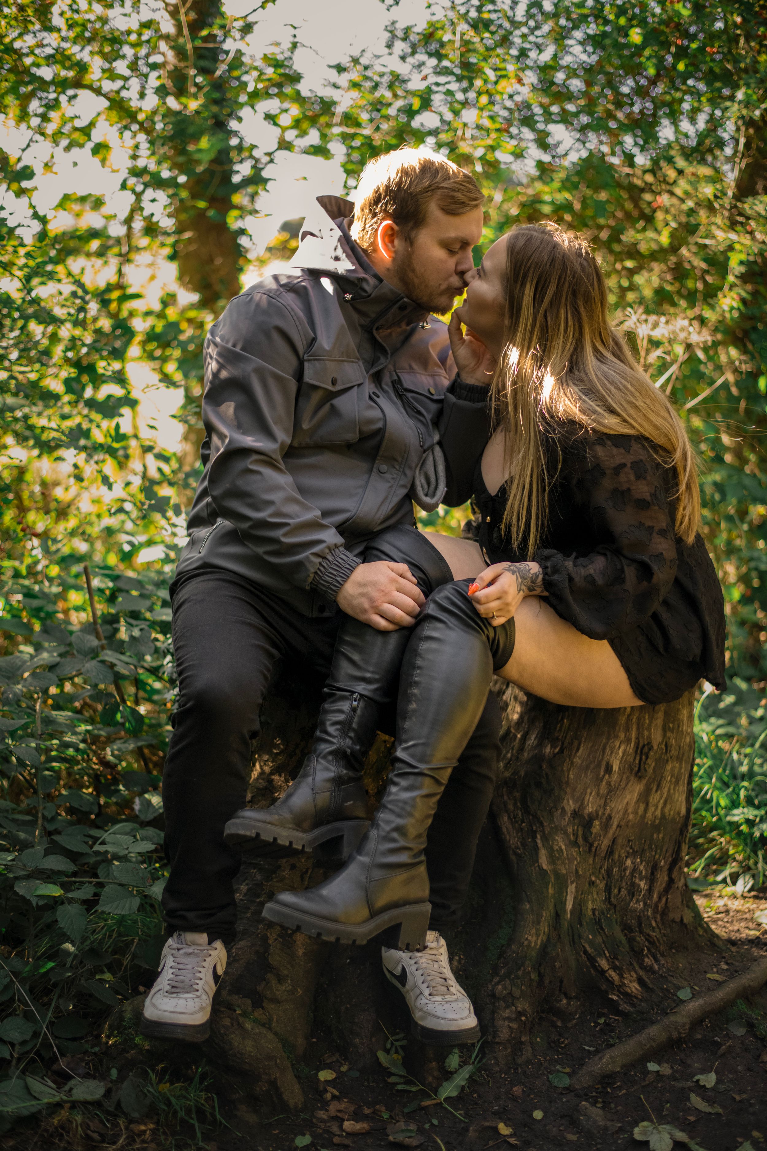 A couple shares an intimate moment while sitting on a tree stump in a sunlit forest.