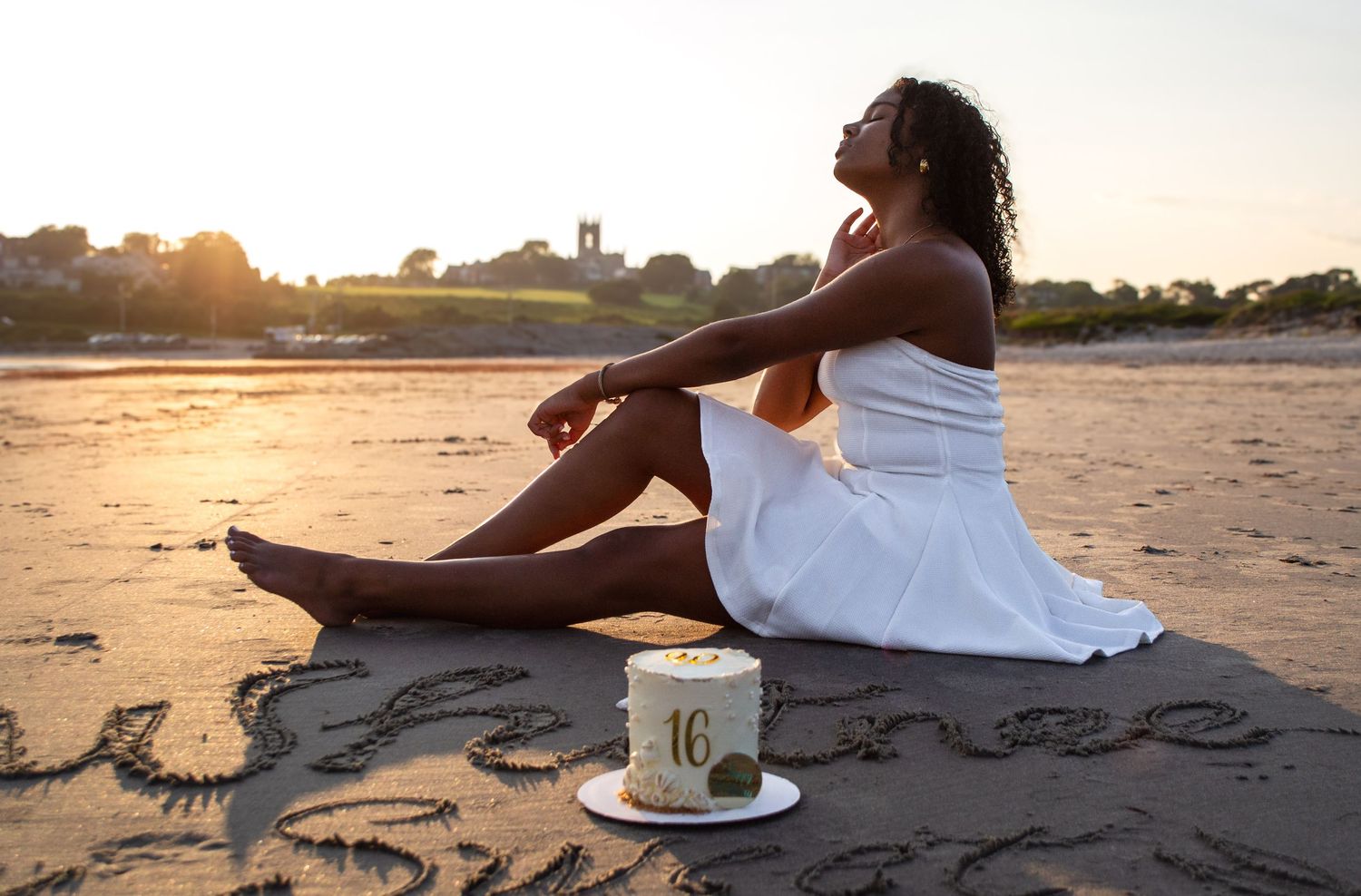 Person in white dress relaxing on sandy beach at sunset with long curly hair and golden lighting.
