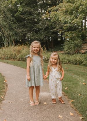 Two young children in summer dresses hold hands while standing on a park pathway.