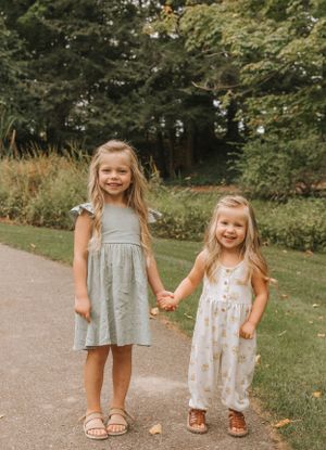 Two children in light-colored dresses smile while standing together on a garden path.