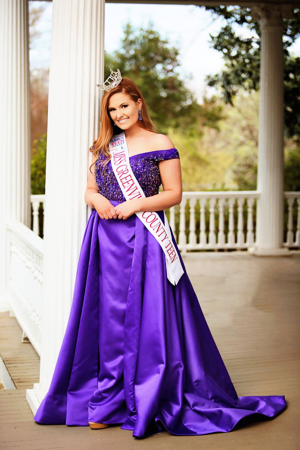 Pageant contestant in purple ball gown and crown posing on white columned porch.