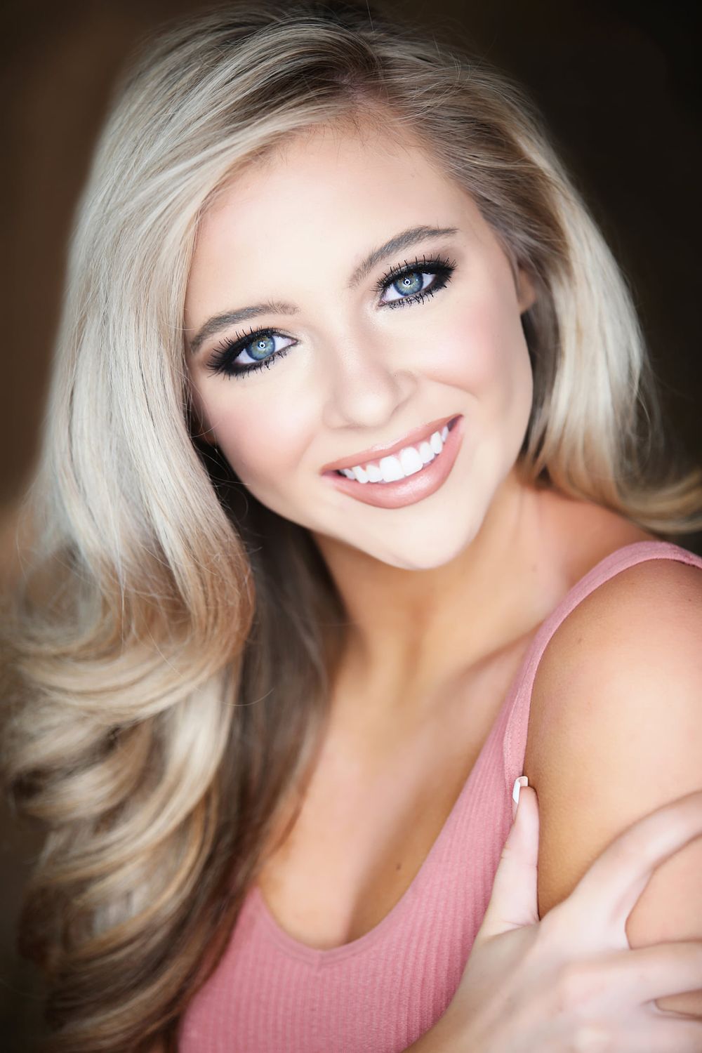Beauty portrait showing bright smile, blonde hair and pink top against dark background.