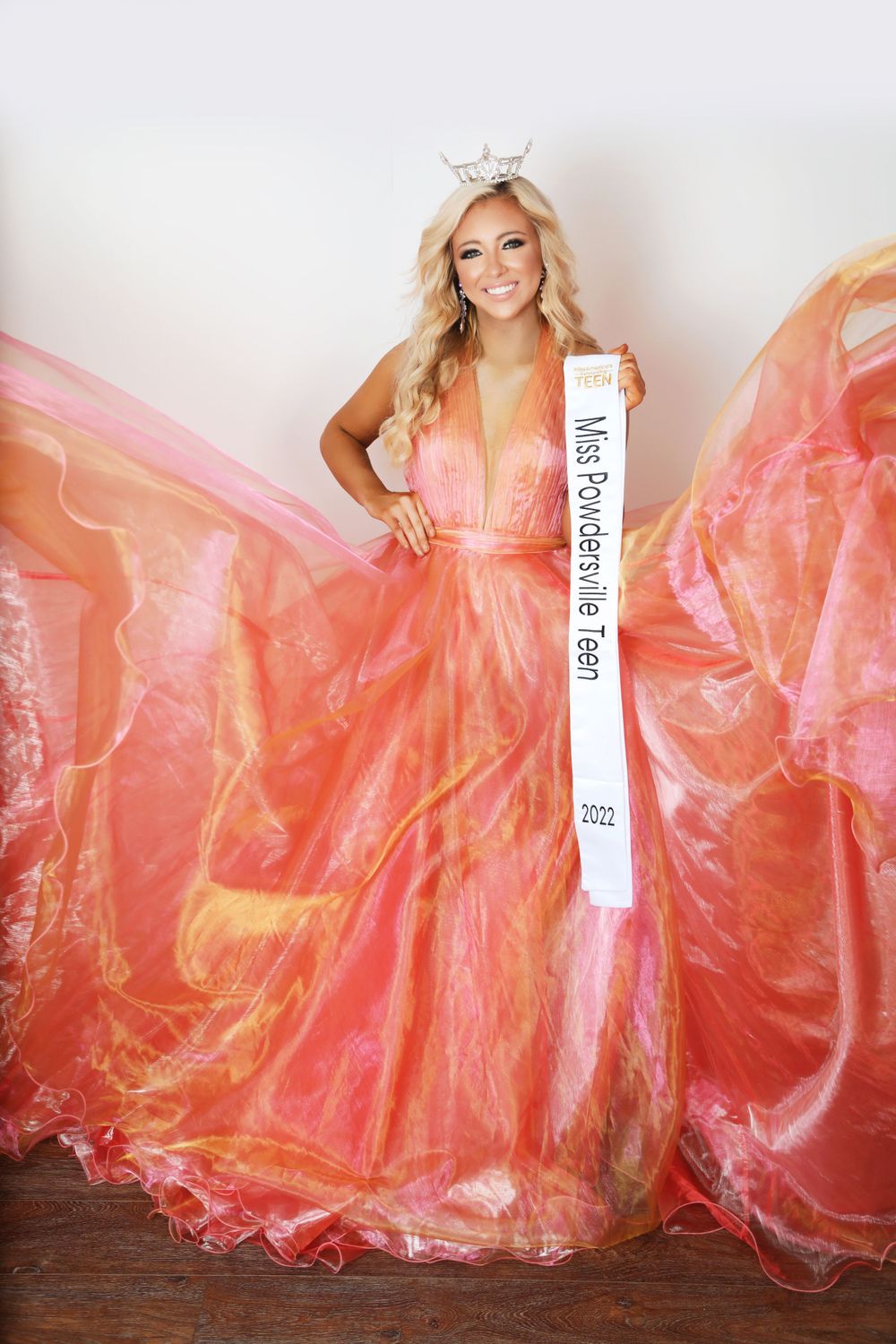 A pageant contestant in a coral gown poses with a white sash and tiara.