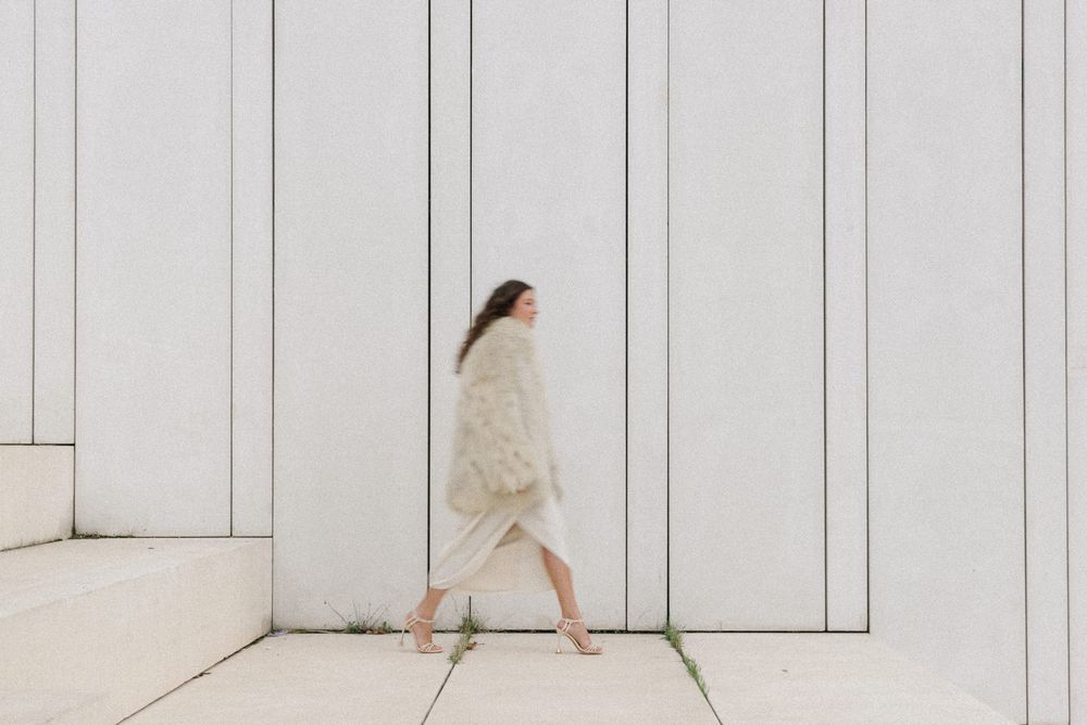 A person in a beige outfit walks along a minimalist white wall with vertical lines, creating a clean architectural scene.