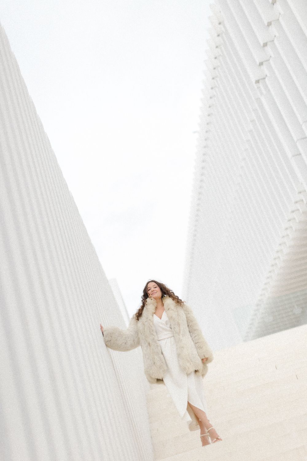 Woman in white winter coat poses dramatically against geometric white architectural backdrop.